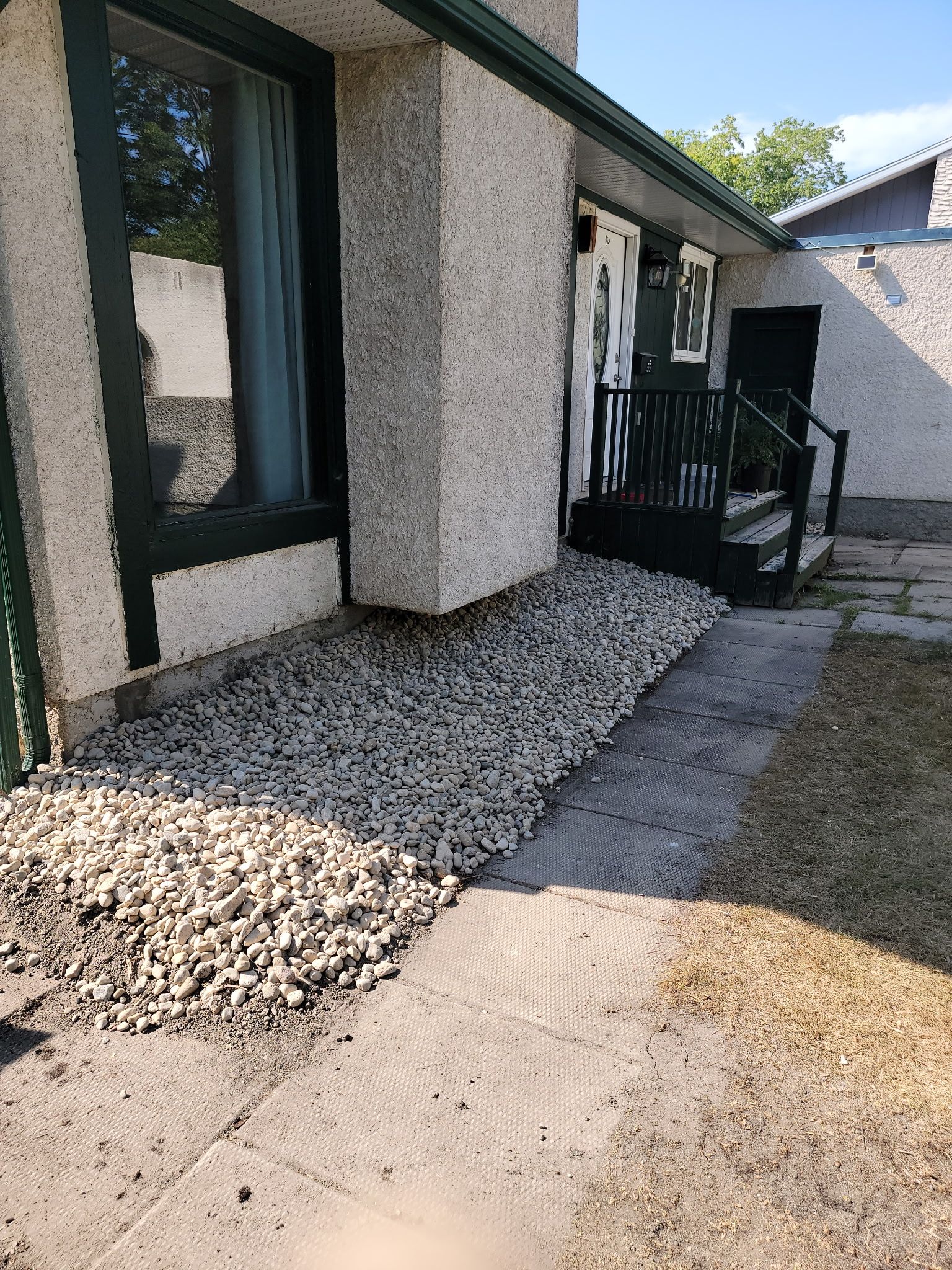 A textured, light-colored house exterior features a dark-framed window, a gravel garden bed, and a small porch staircase.