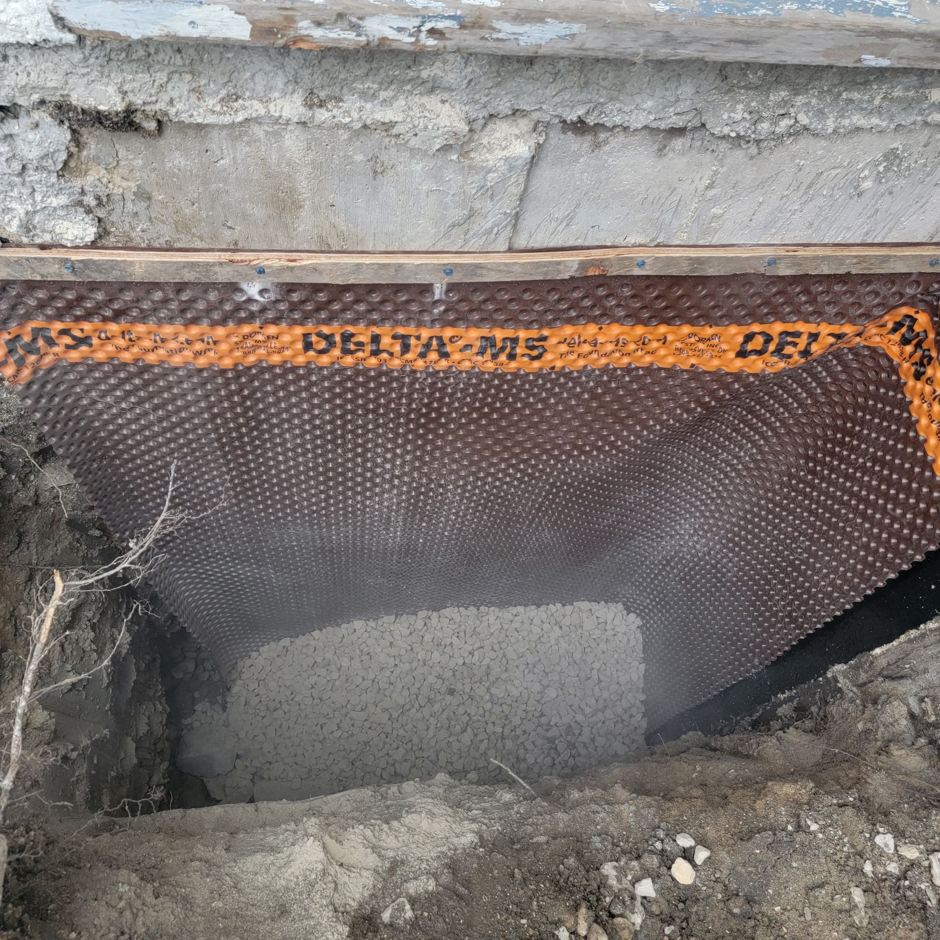 Foundation wall with brown DELTA-MS dimpled drainage membrane installed against concrete, surrounded by excavated dirt.