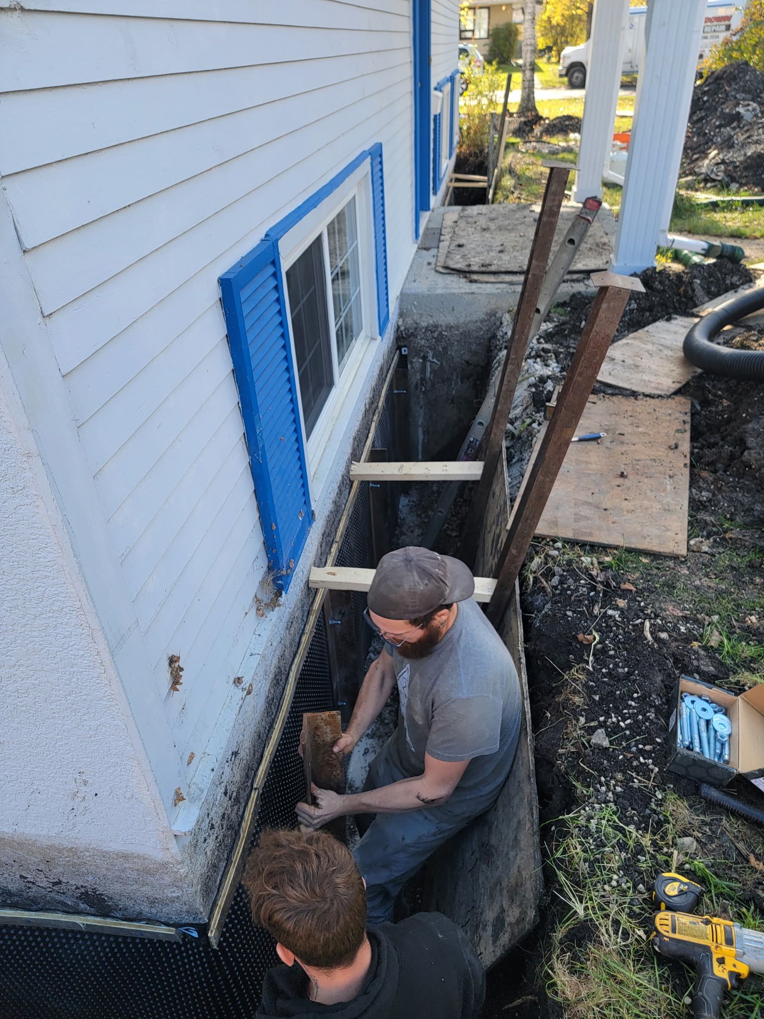 Two workers installing foundation waterproofing materials in a deep trench along the side of a white house.