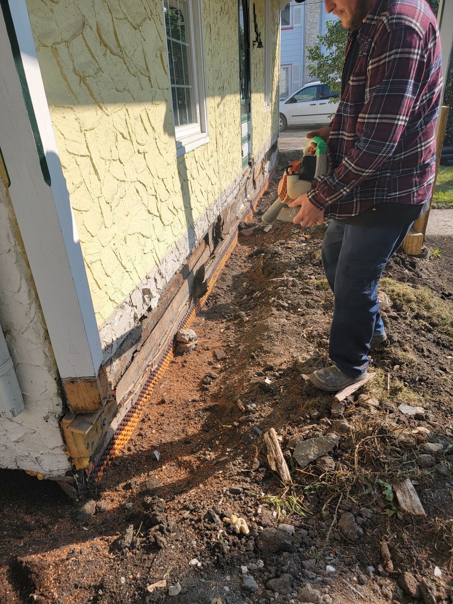 A person in a plaid shirt uses a handheld power tool to cut into the wood siding near the foundation of a yellow house.
