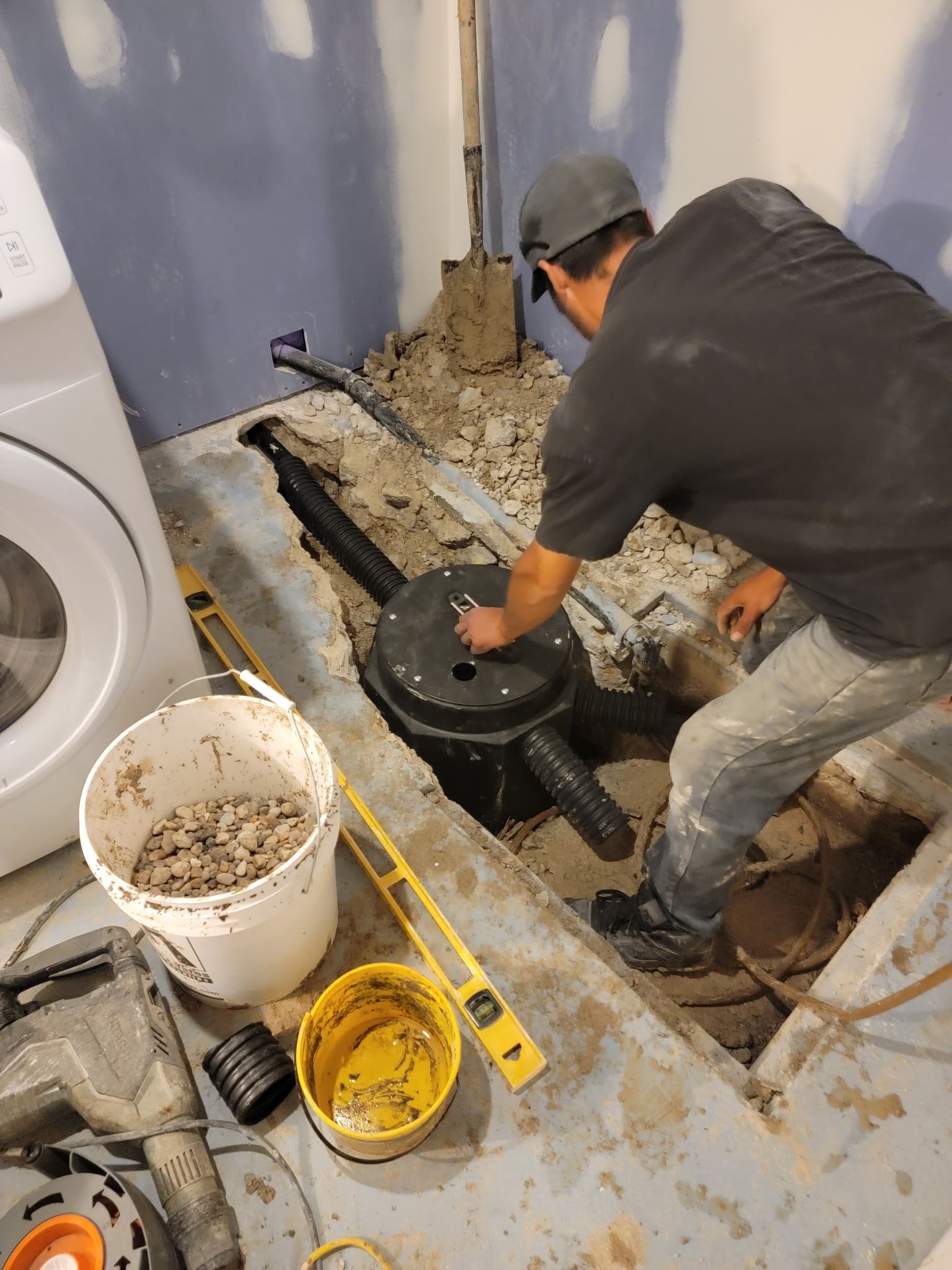 A person kneeling on a basement floor repairs a black sump pump basin installed in a trench dug into the concrete.