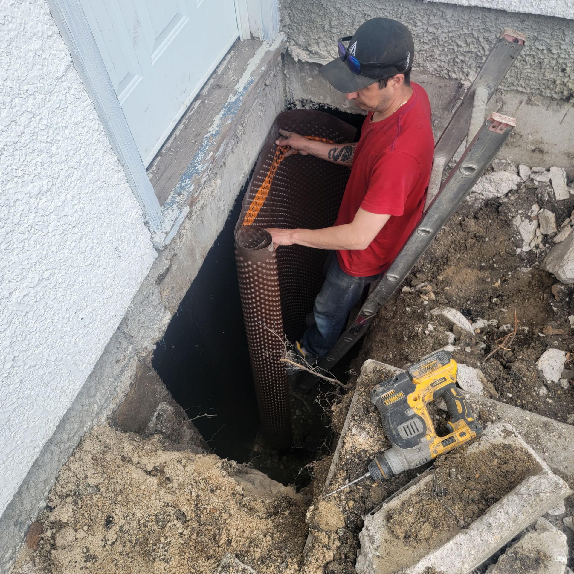 A person in a red shirt installs a dimpled foundation drainage membrane along an exterior wall in an excavated trench.