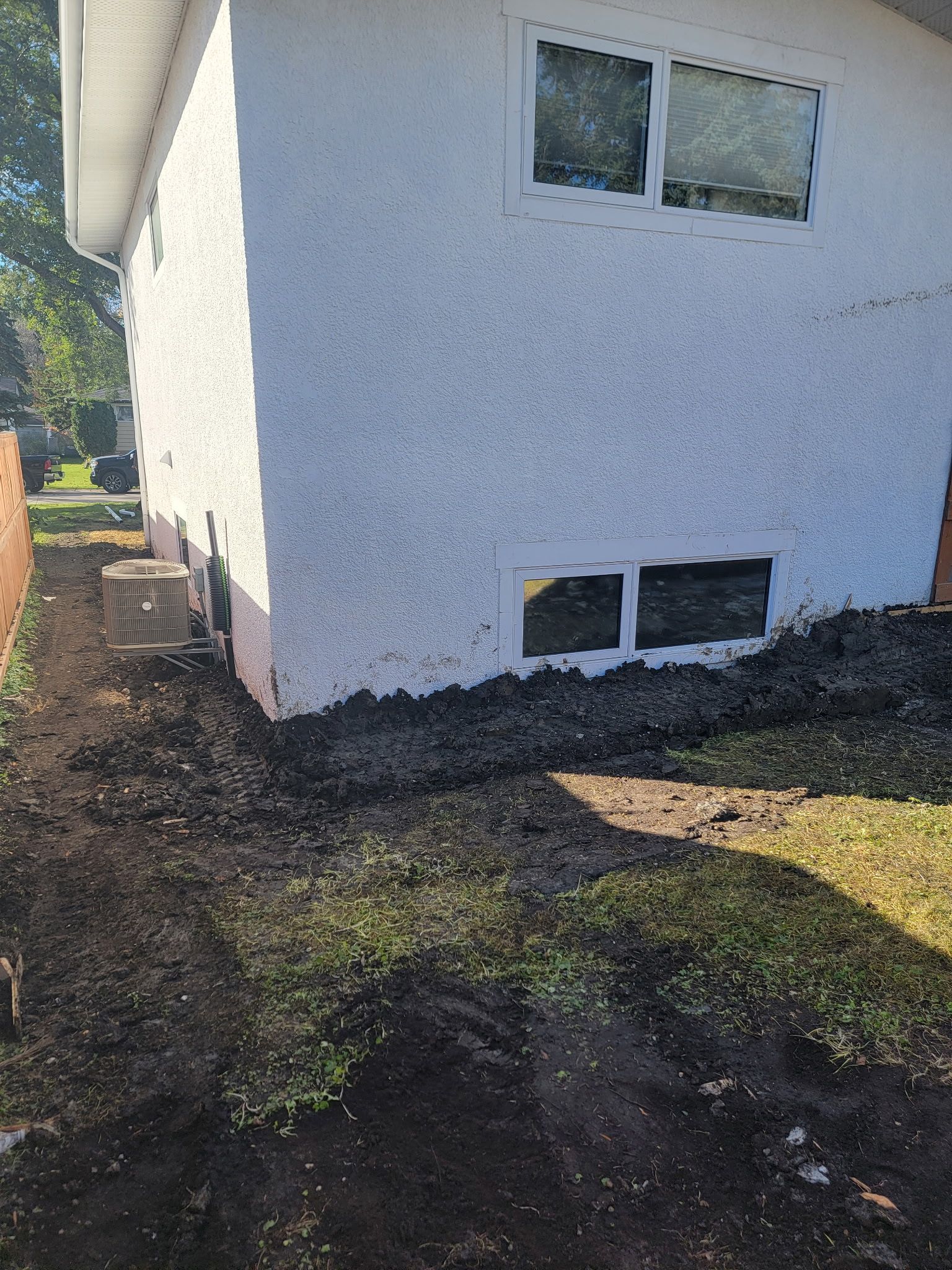 A side view of a white, textured house exterior with two windows and a dark, newly excavated dirt area along the foundation.
