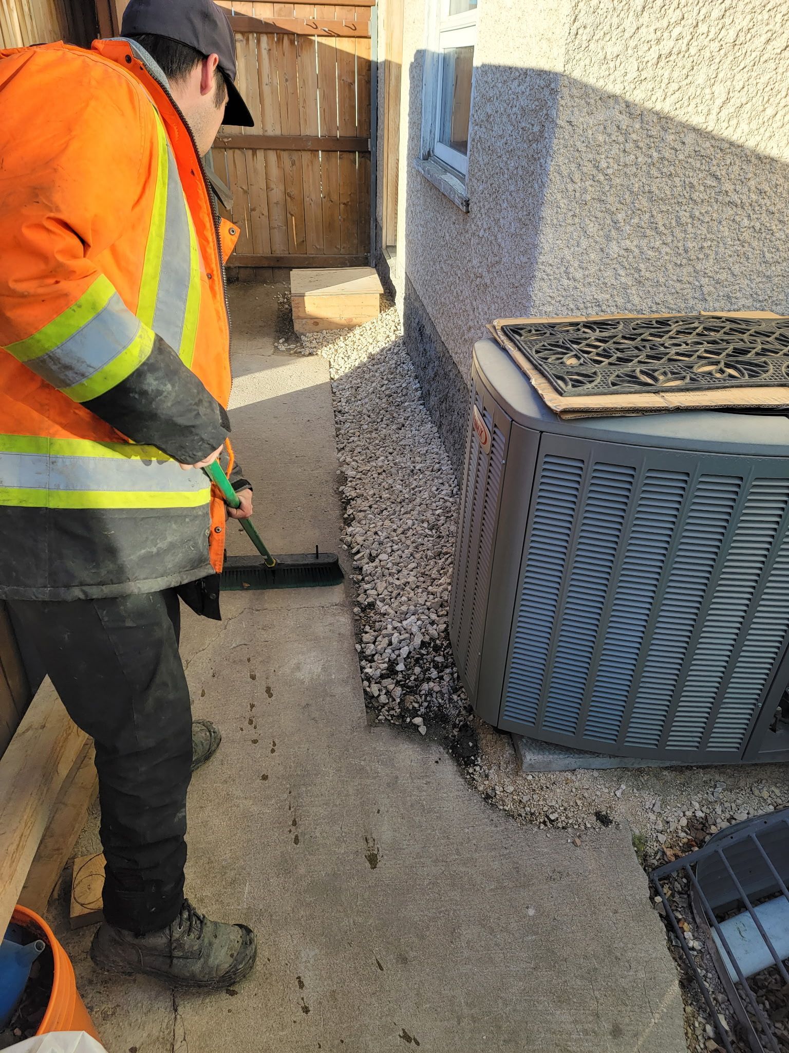 A person in a high-visibility orange jacket uses a broom to sweep gravel next to an outdoor HVAC unit against a house.