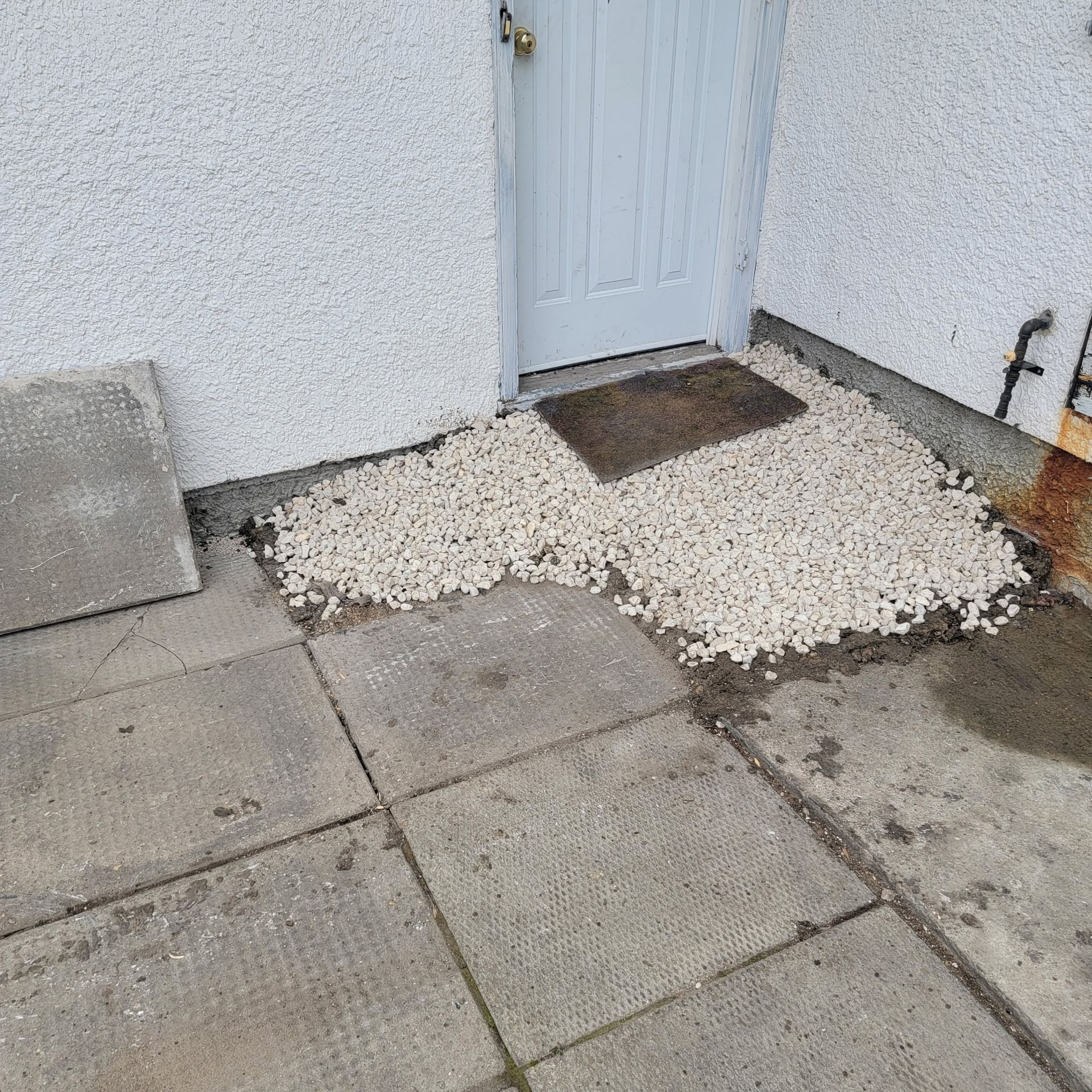 A white exterior wall and door with a patch of gravel, a dark doormat, and gray concrete paving slabs on the ground.
