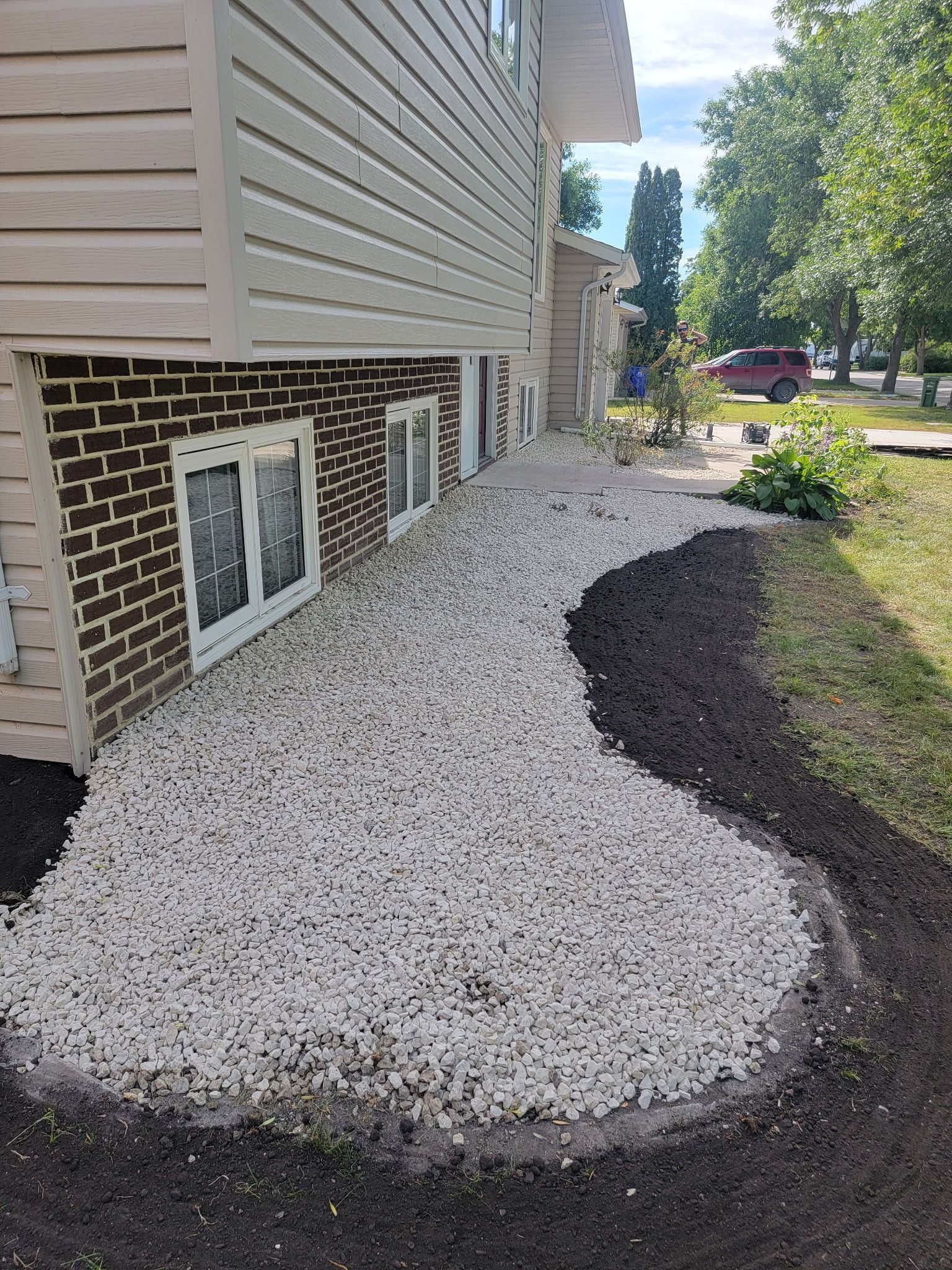 A tan house exterior with a basement brick wall and windows above a new decorative gravel garden bed with mulch edging.