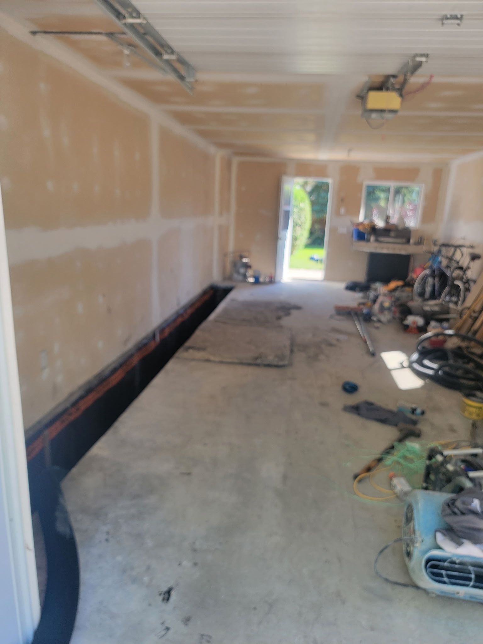 A dimly lit garage interior under construction with drywall-covered walls and miscellaneous tools and gear on the floor.