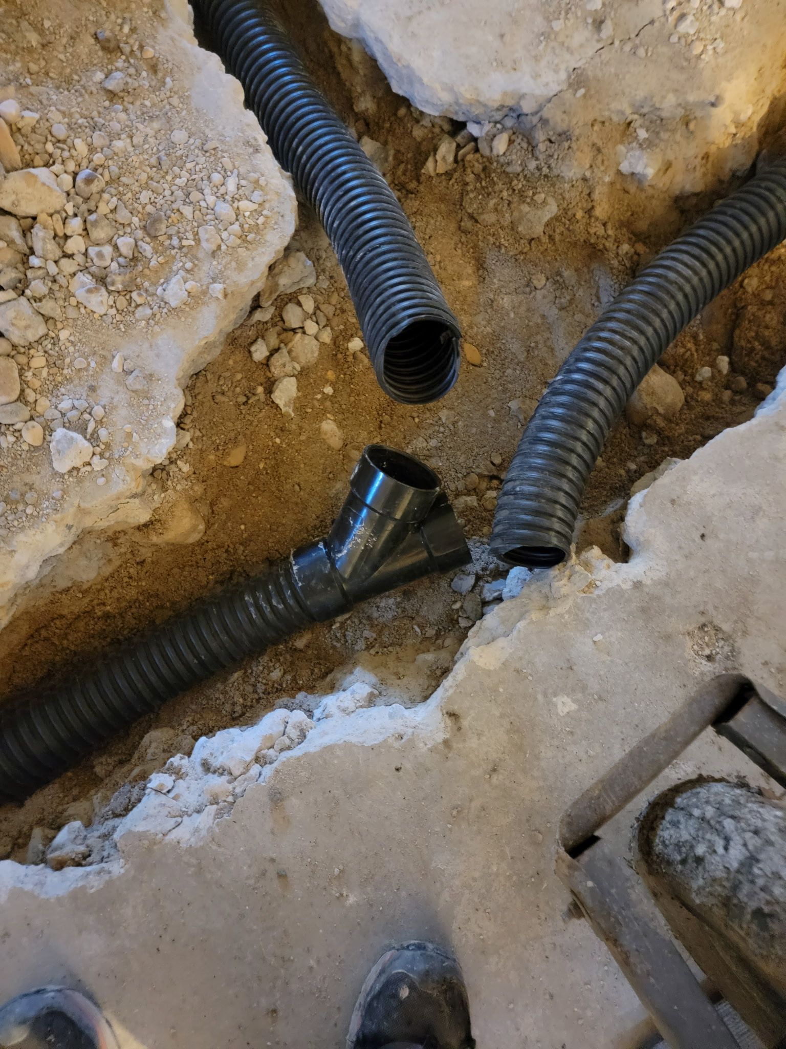 Black flexible drainage pipes are positioned in a dirt-filled trench cut through a concrete floor.