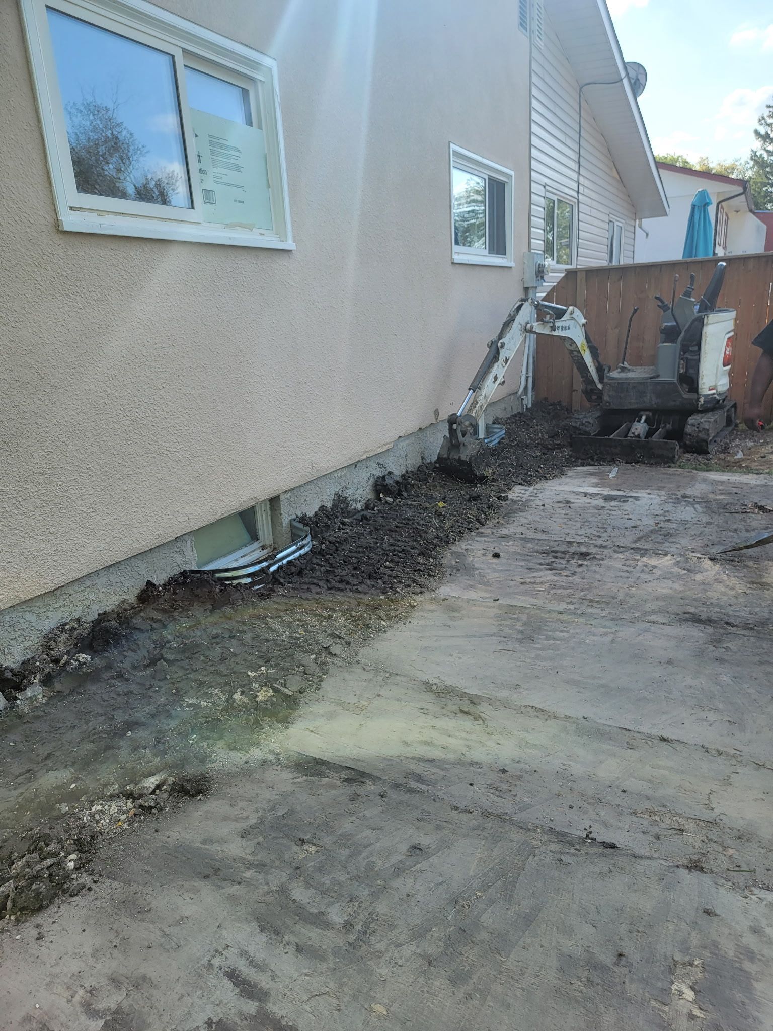 A small excavator digs a trench along the concrete foundation of a beige house beside a gravel driveway.