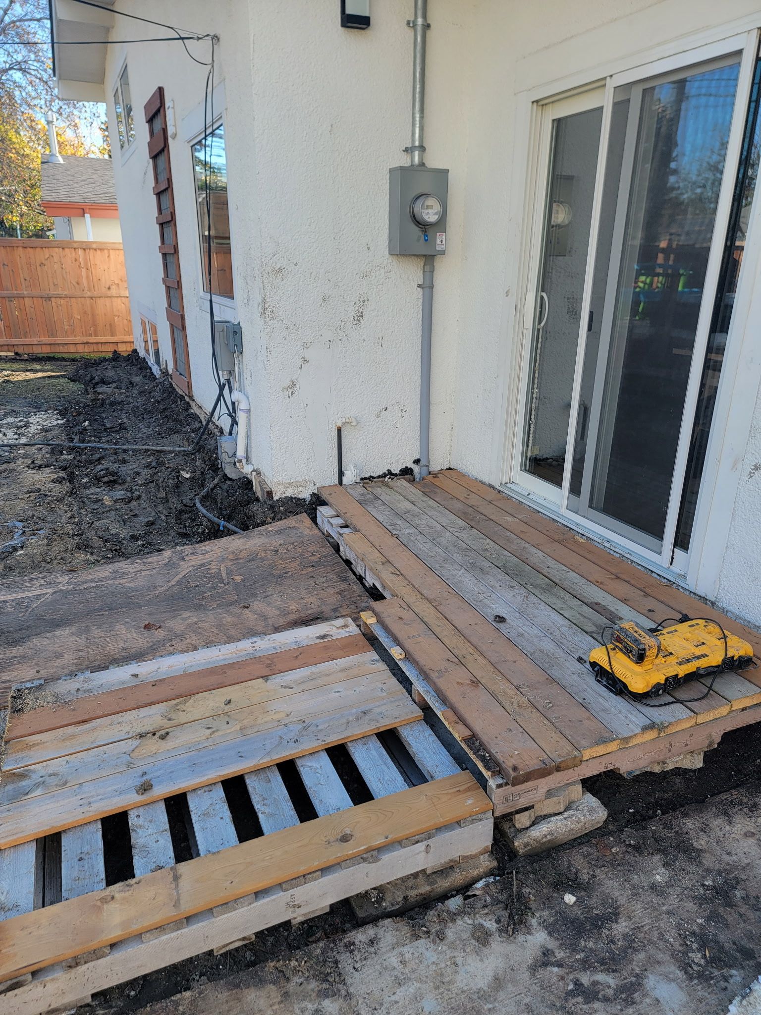 A wooden pallet deck under construction outside a white stucco house, next to a sliding glass door and a power meter.