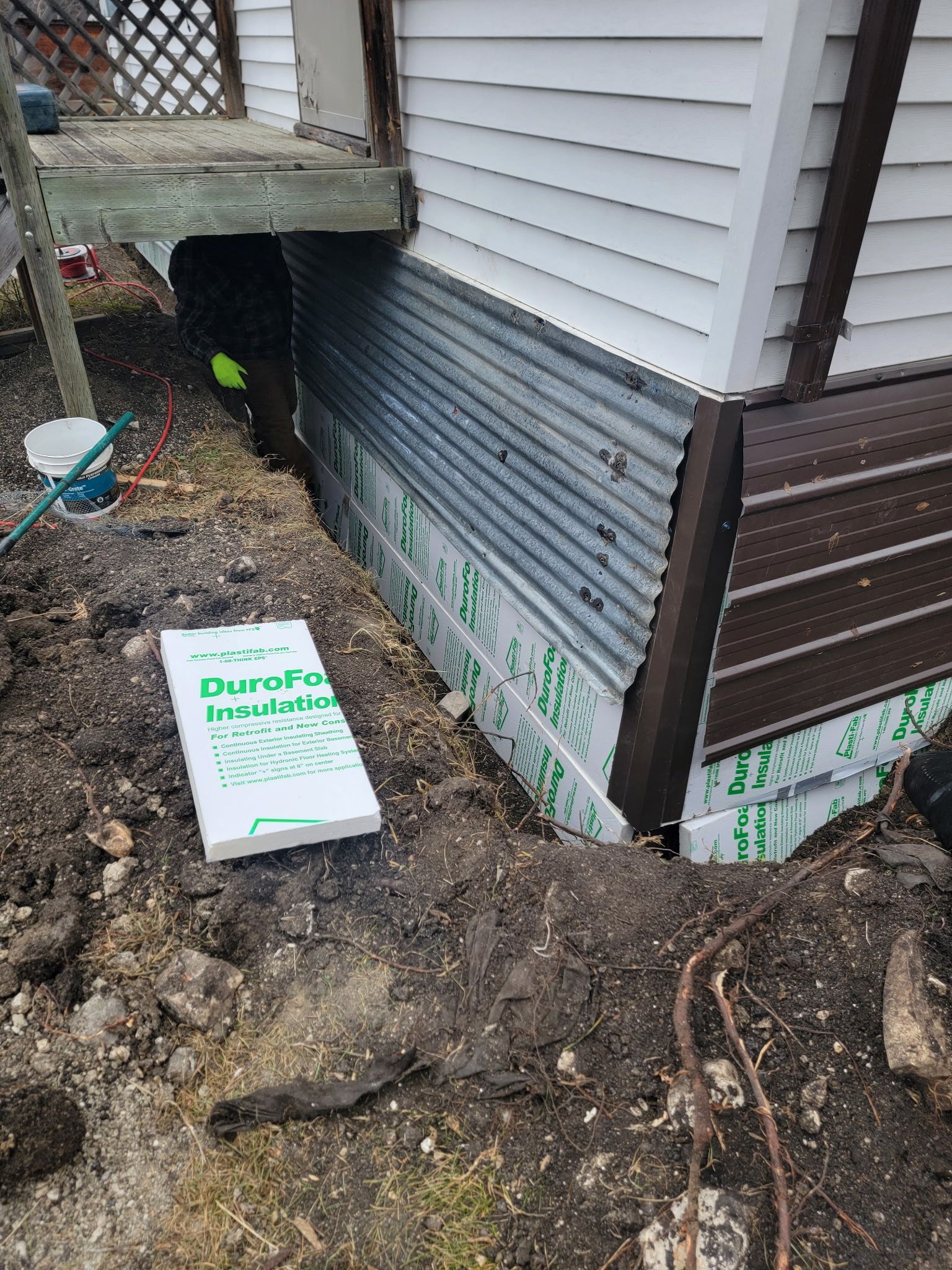 Exterior house foundation with corrugated metal and insulation panels visible above dirt, near a concrete porch step.