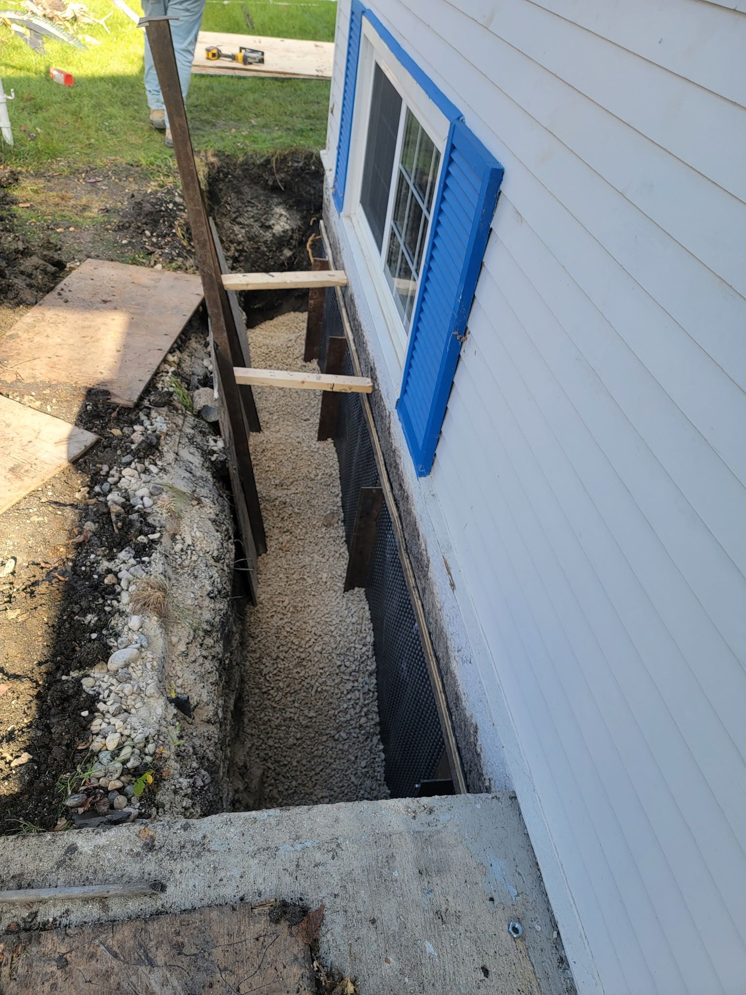 A trench has been dug along the side of a white house with blue shutters, exposing the foundation covered by black mesh.