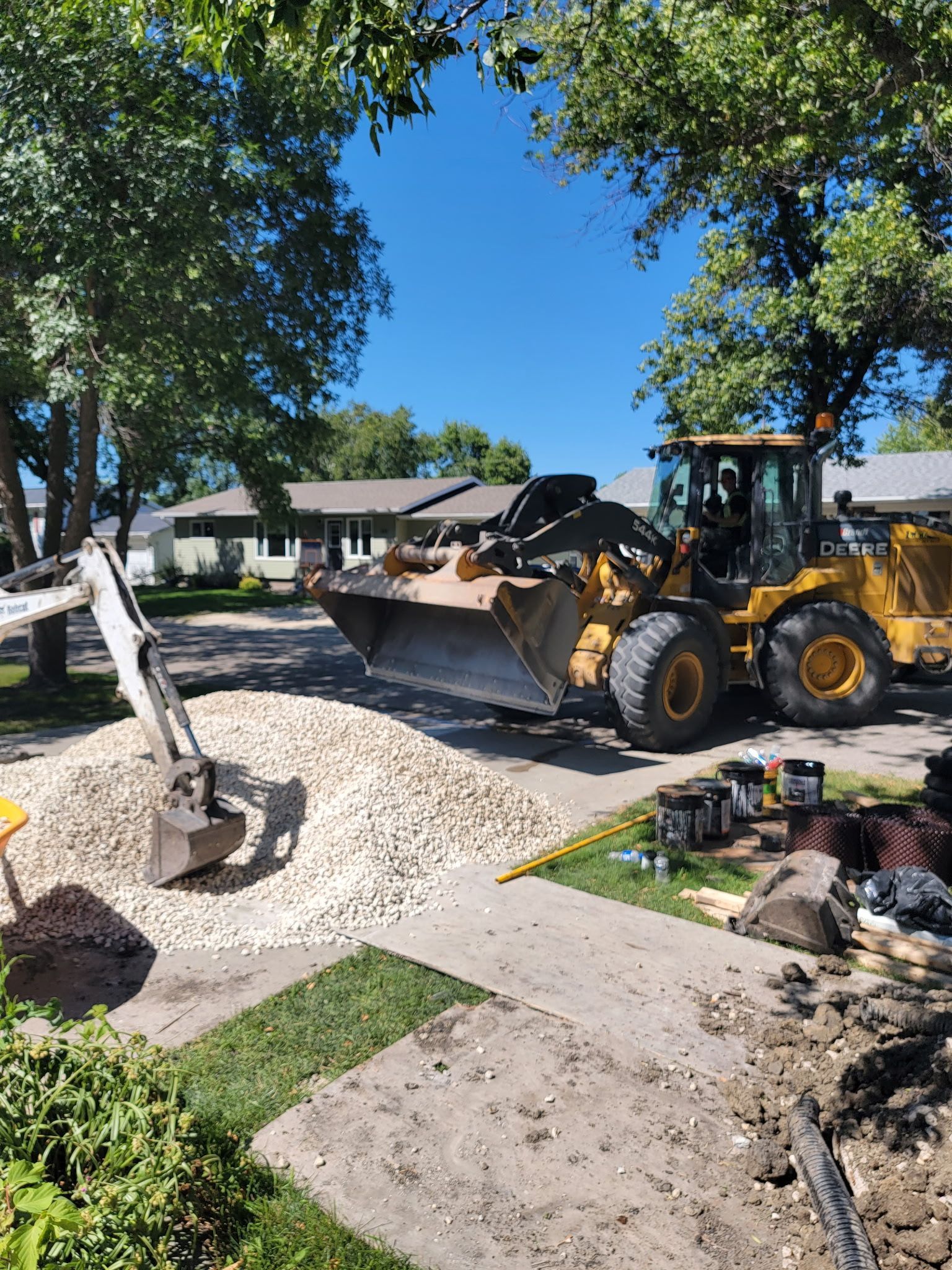 A yellow front-end loader and a small excavator move a large pile of gravel on a residential driveway on a sunny day.