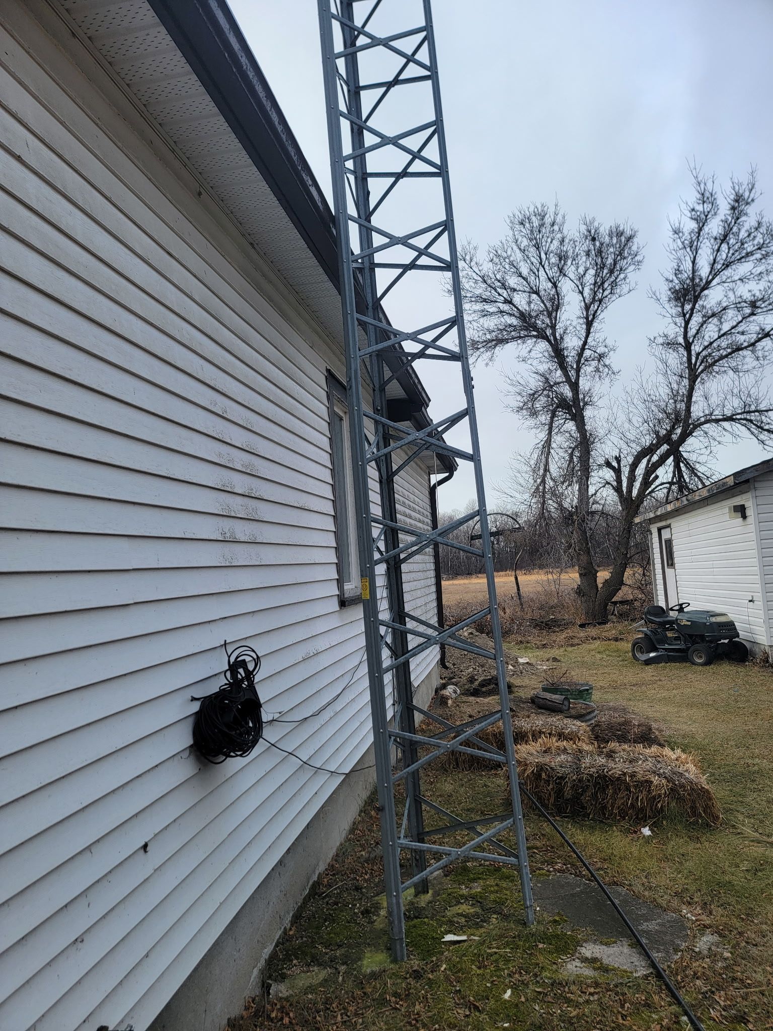 A tall, gray metal communication tower stands against the white-sided exterior wall of a house near a yard.