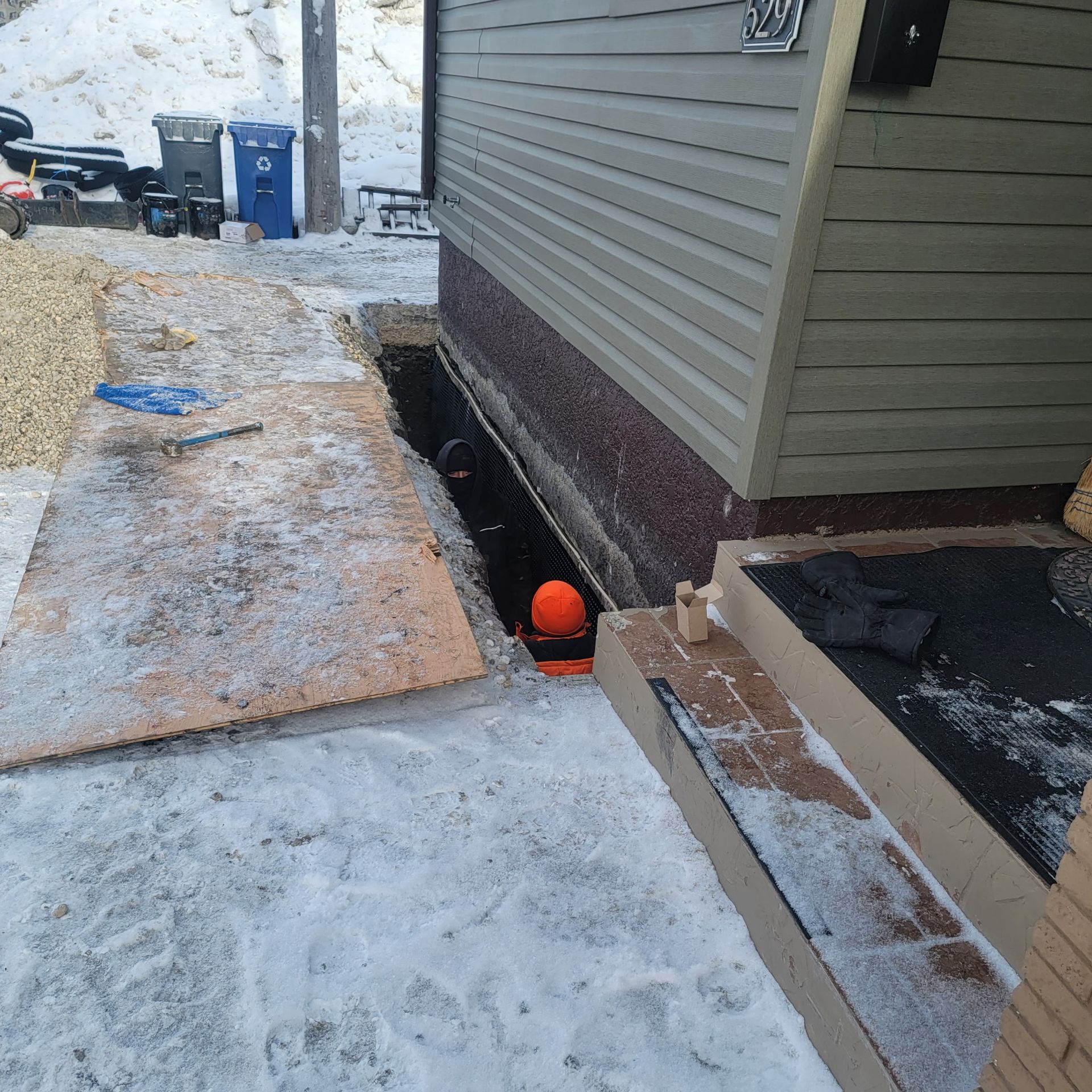 A worker in an orange hard hat is inside a narrow trench dug along the foundation of a house during winter.