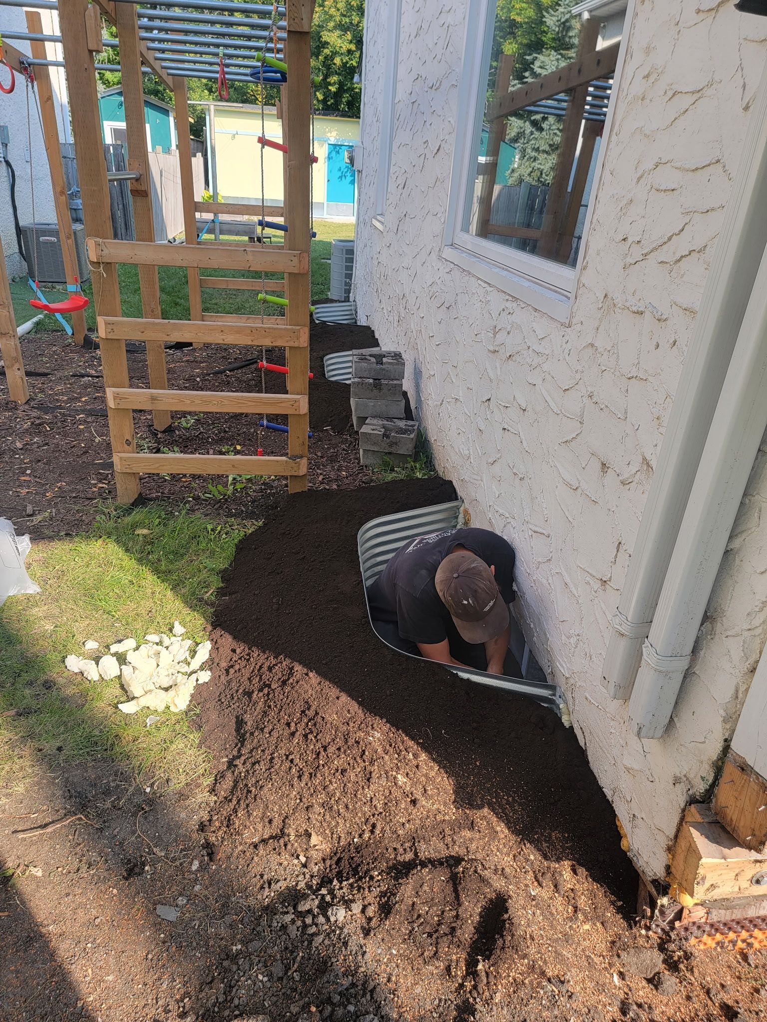 A person installs a corrugated metal window well against a stucco house exterior in a mulched yard.