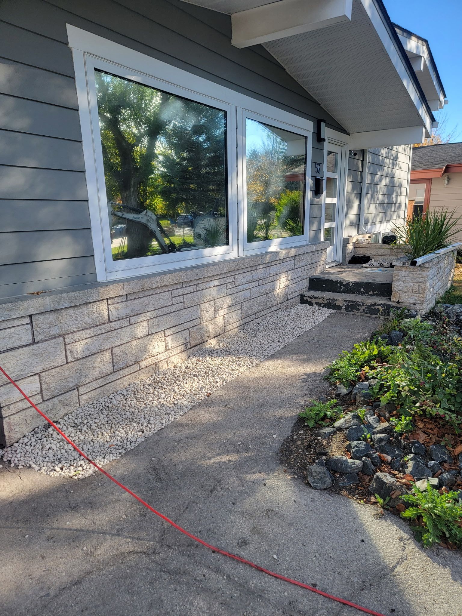 A grey house exterior featuring a large window, a light-colored brick foundation, and a concrete walkway.
