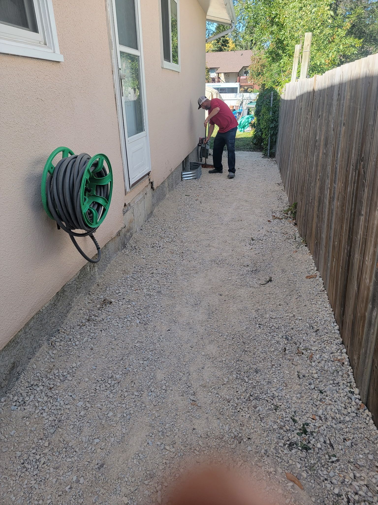 A person in a red shirt works near the foundation of a house next to a gravel path and a wooden fence.