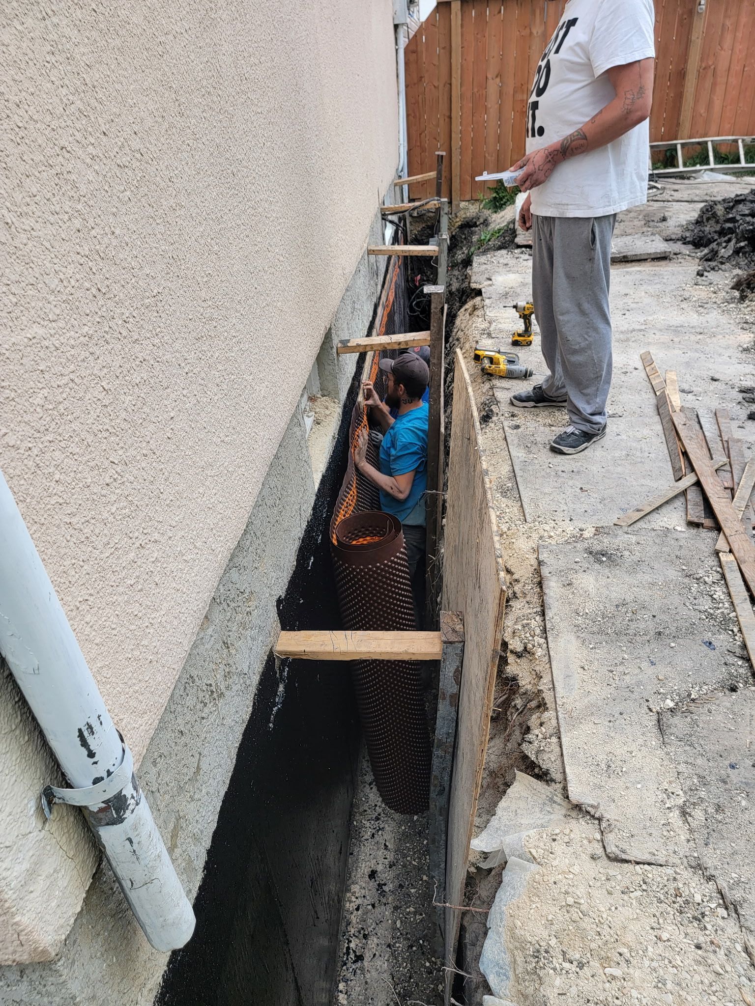 A person works in a trench beside a building foundation with protective drainage sheeting and wooden support bracing.