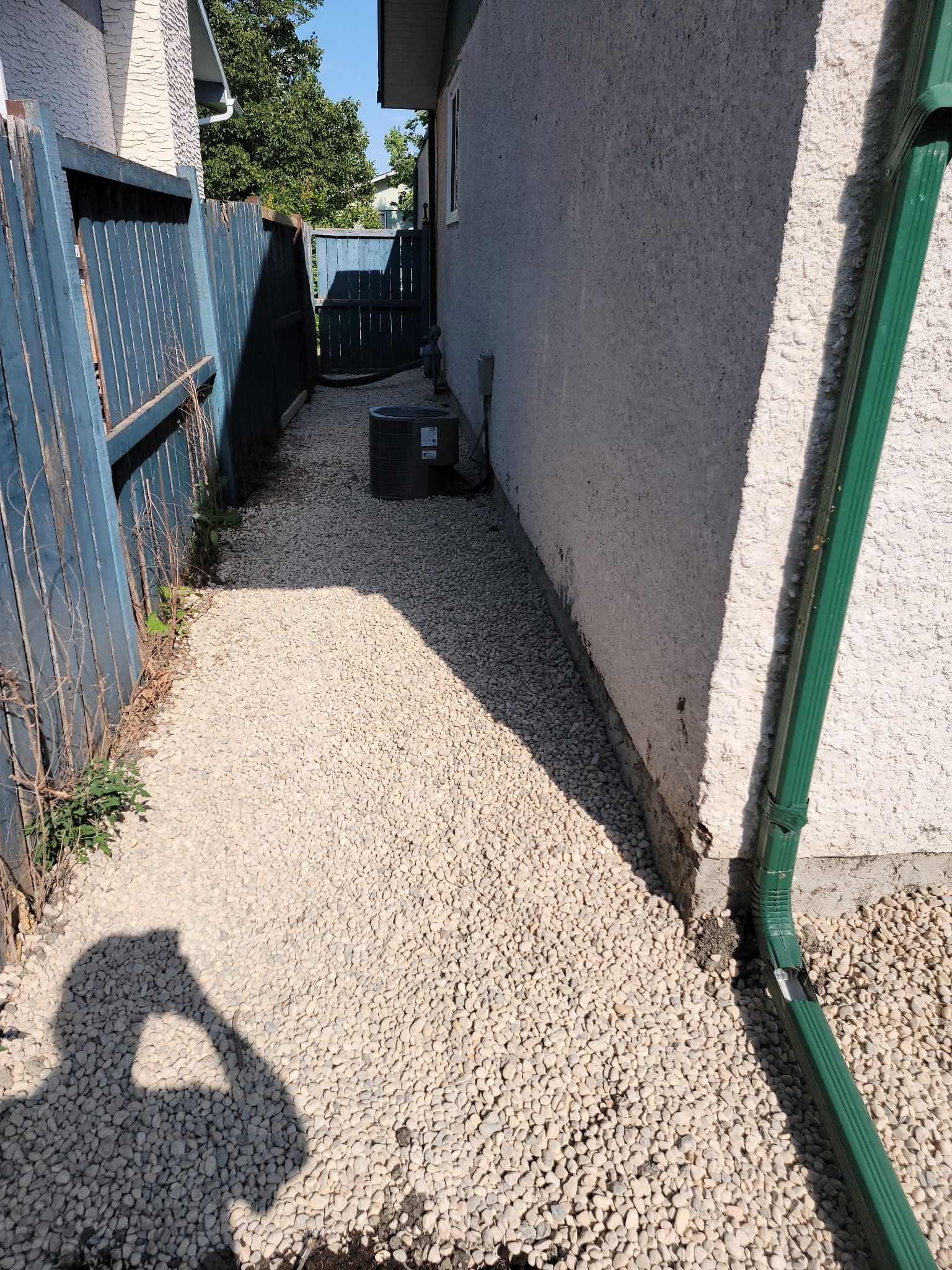 A gravel side yard beside a stucco house with a blue fence on the left and a green downspout on the right.