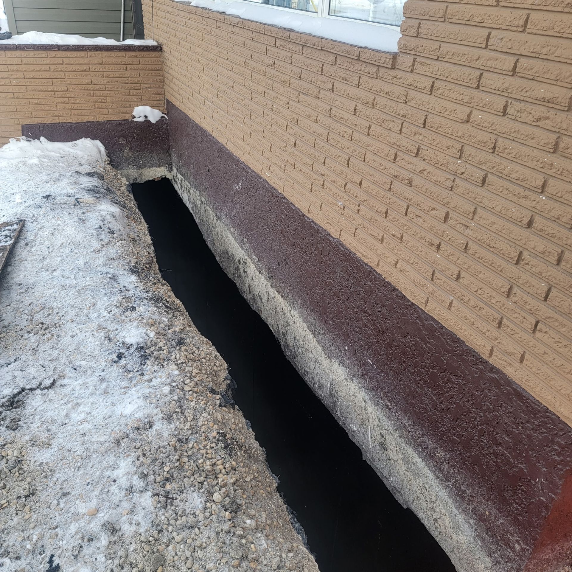 A trench dug along the exterior foundation of a brick house, exposing a section of the basement wall.