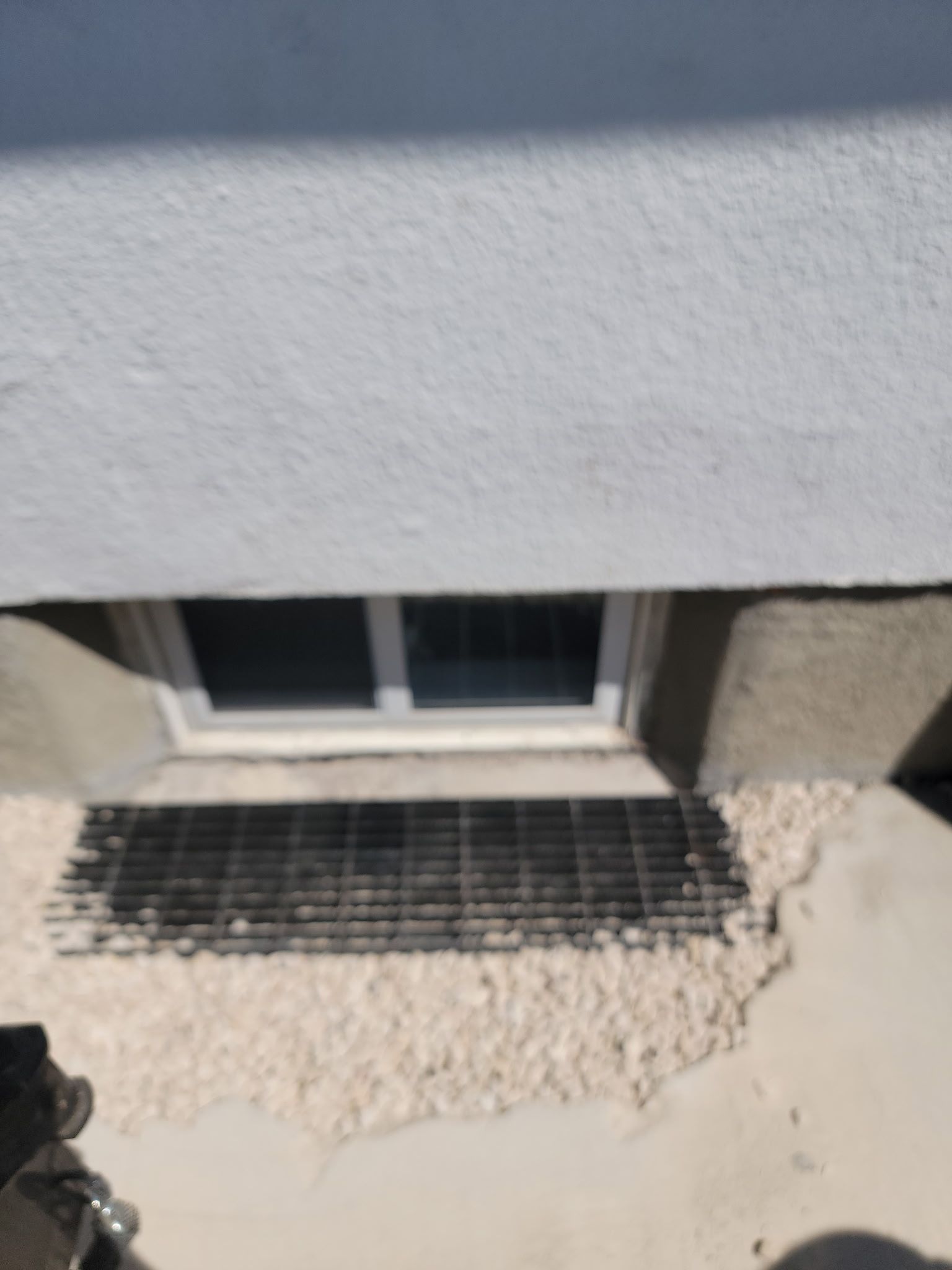 A basement window at ground level with a metal safety grate covering a gravel window well against a white exterior wall.