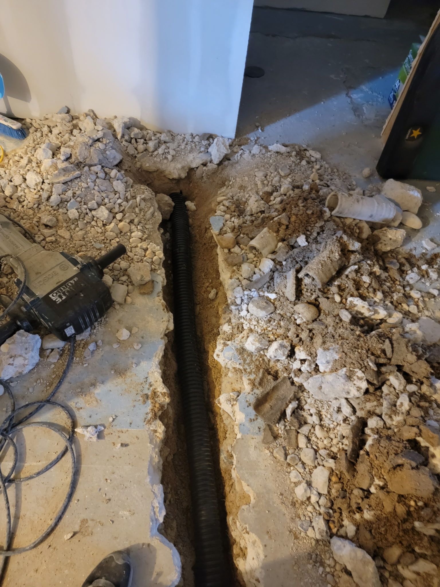 A black corrugated pipe lies in a trench cut through a concrete floor, surrounded by piles of rubble and concrete debris.