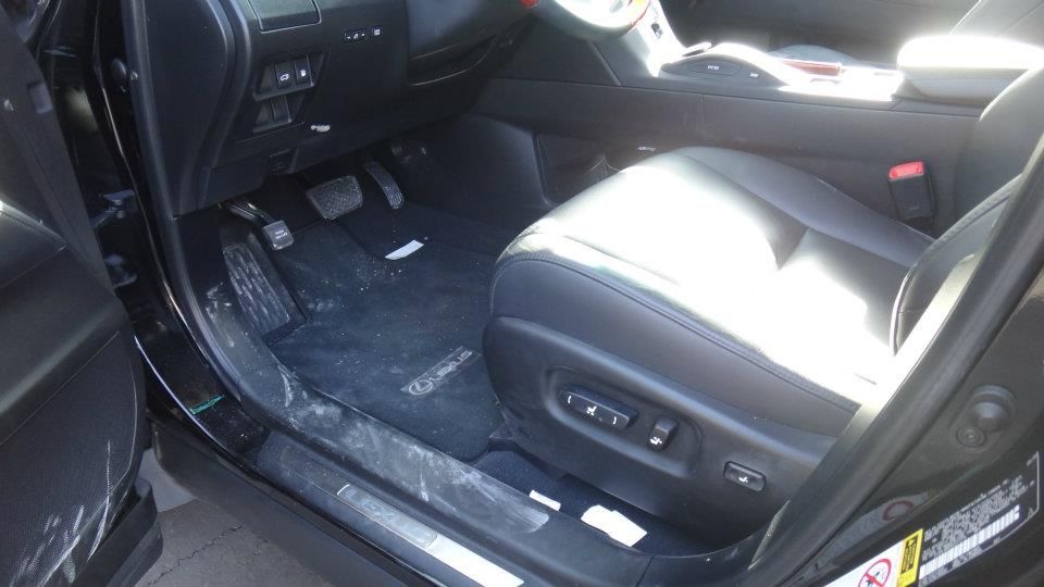 Close-up of a black car's interior driver-side footwell and leather seat showing dust and debris before a professional interior detailing.