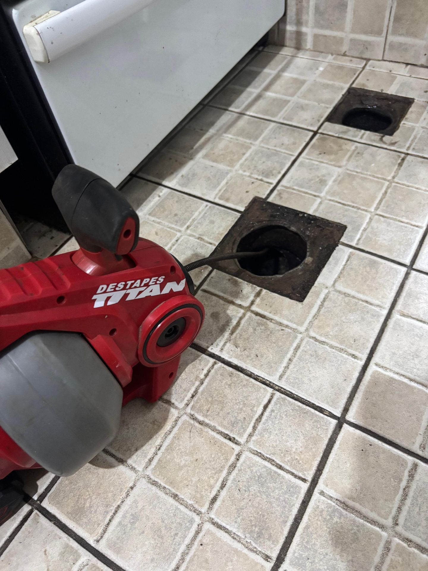 Una desatascadora roja limpia un desagüe en un suelo de baldosas. Se ve un electrodoméstico blanco.