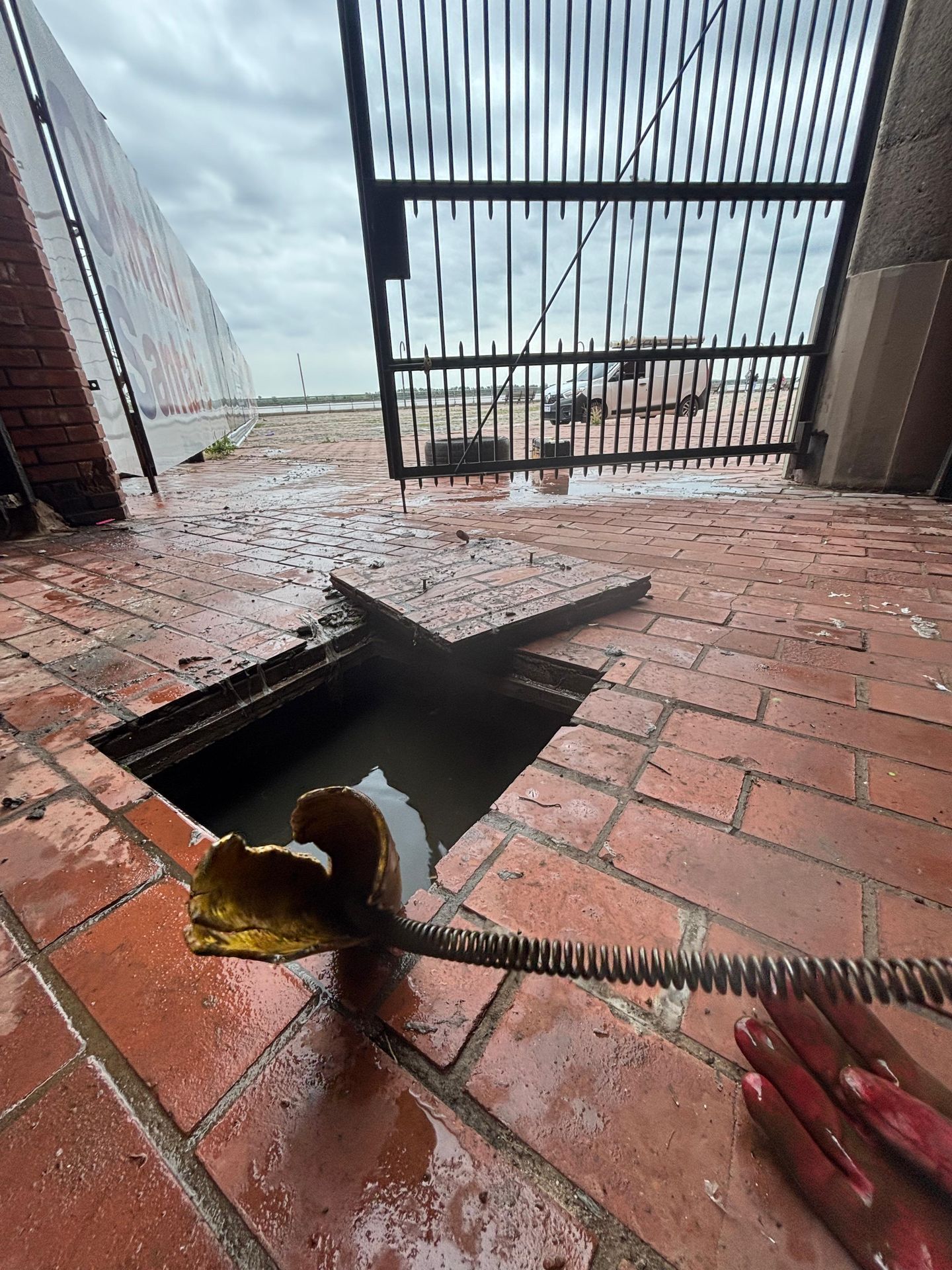 Acceso abierto al alcantarillado con agua, serpiente de drenaje y patio de ladrillos mojados; puerta en el fondo.