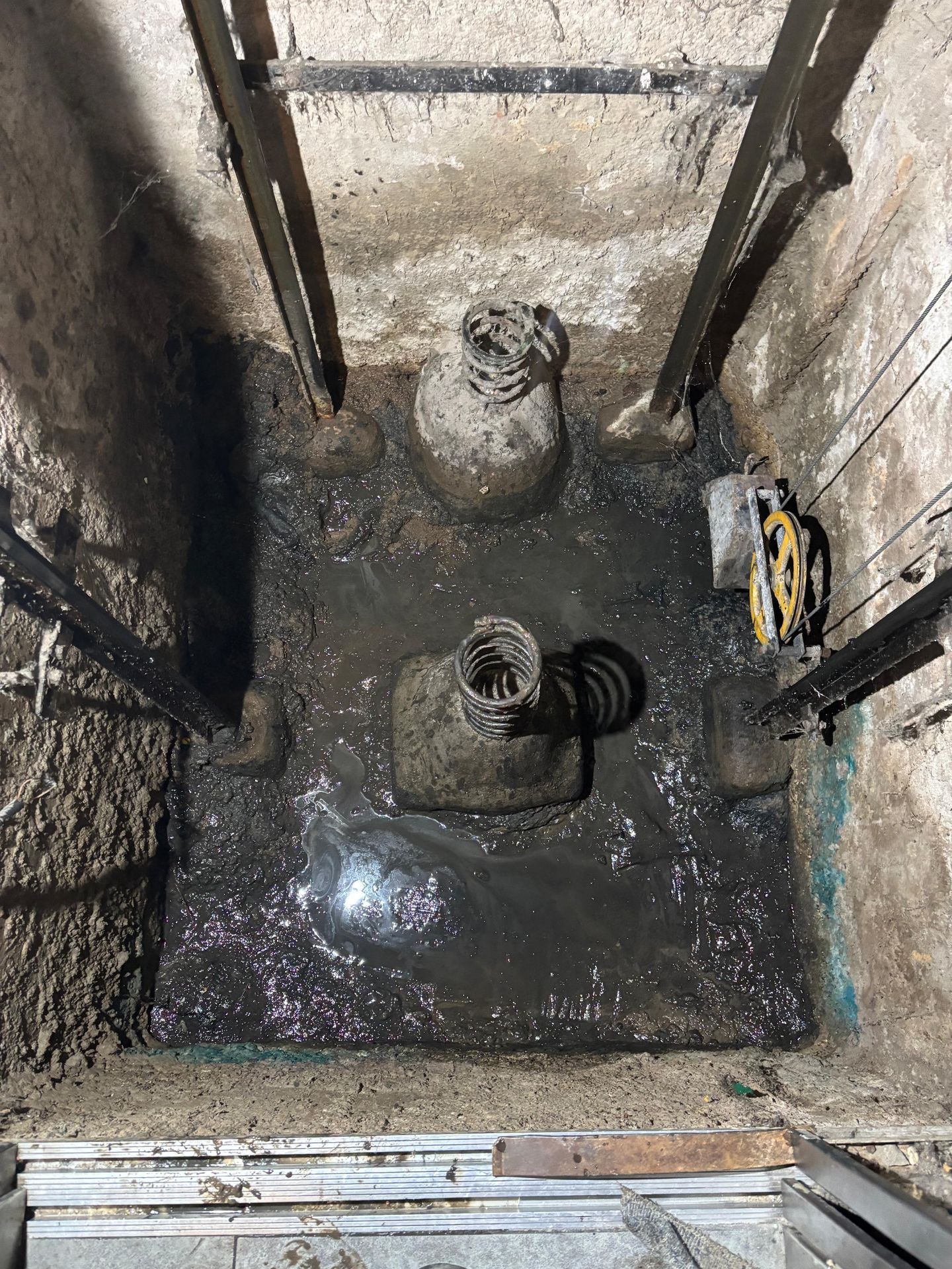 Mirando hacia abajo a un pozo de hormigón húmedo y oscuro con dos tuberías y vigas metálicas de soporte.