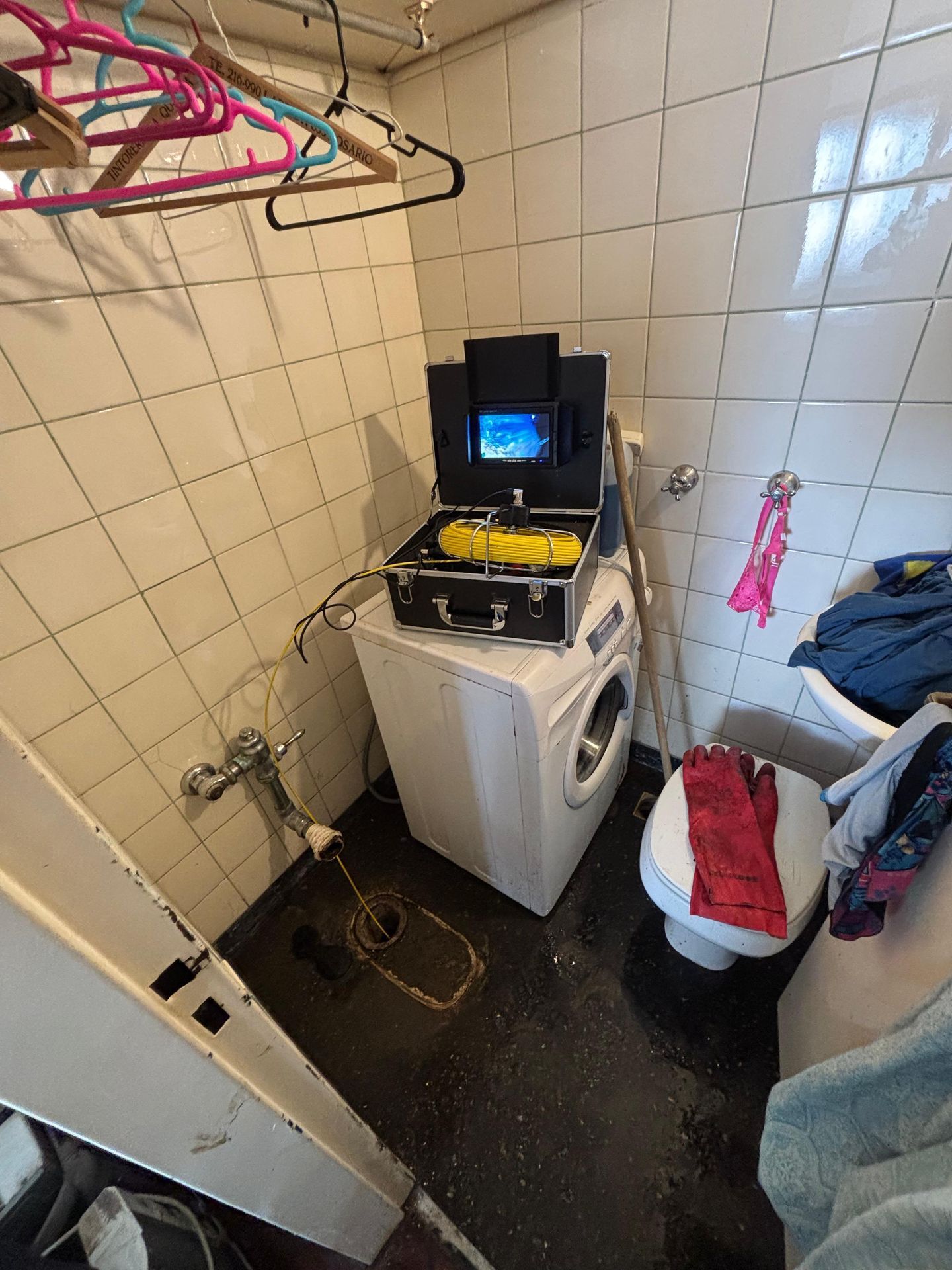 Equipamiento de una lavadora en un baño pequeño. Caja negra con monitor; mangueras amarillas. Ropa, inodoro y azulejos.