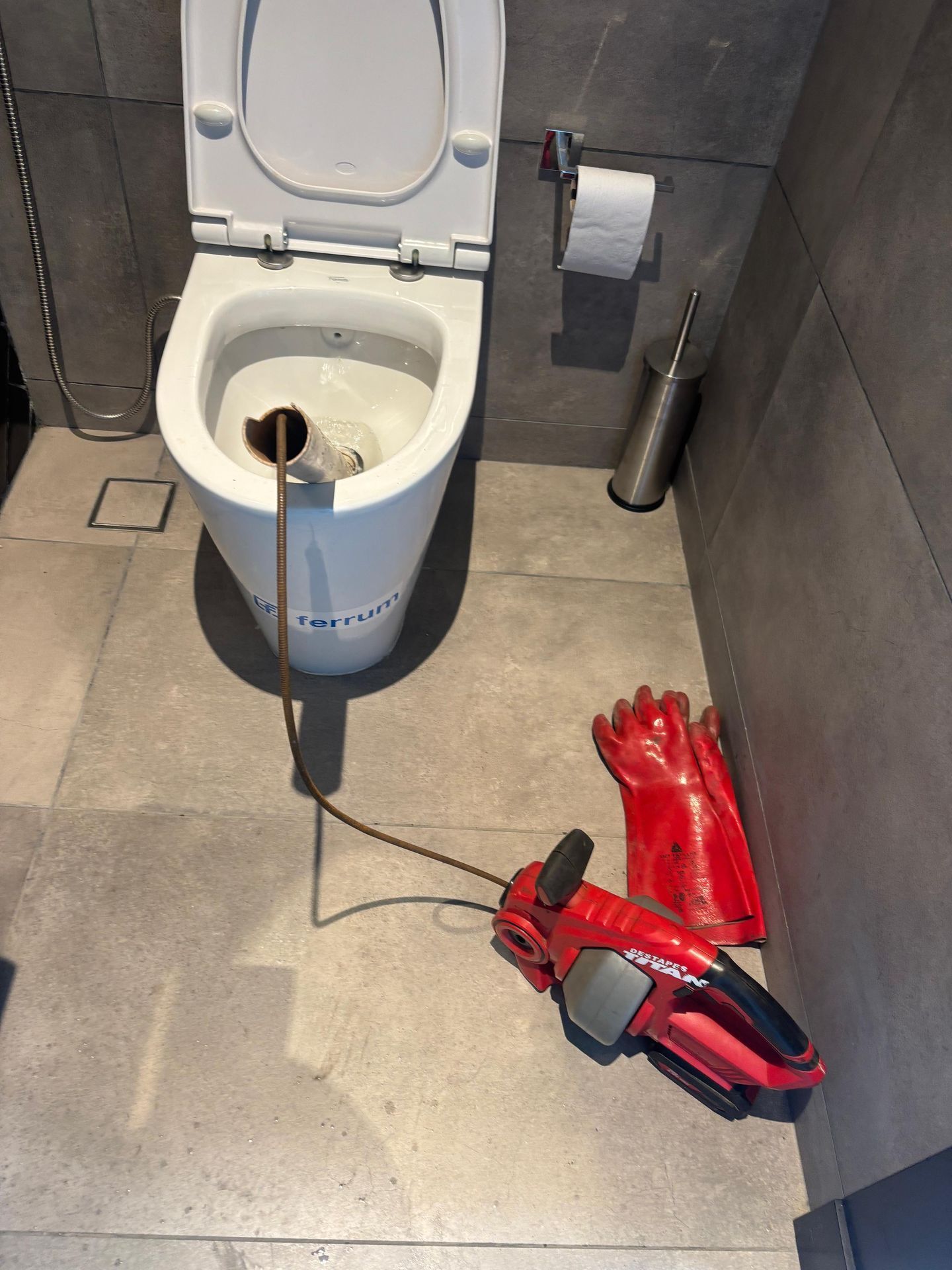 Inodoro con desatascador en su interior, junto a guantes y una herramienta eléctrica. Baño.