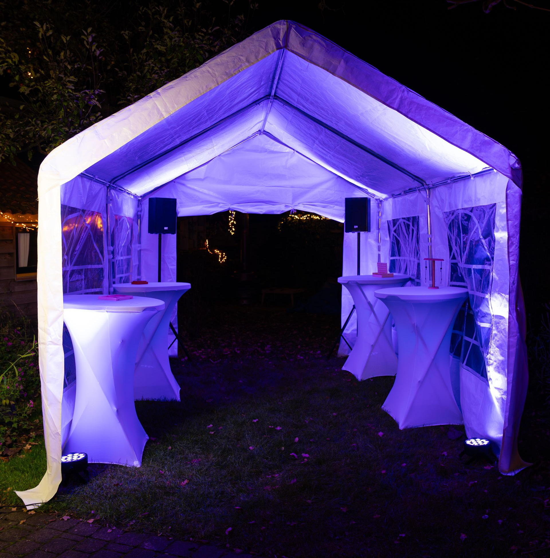 Een verlichte witte buitentent 's nachts met vier cocktailtafels, paarse sfeerverlichting en twee luidsprekers.