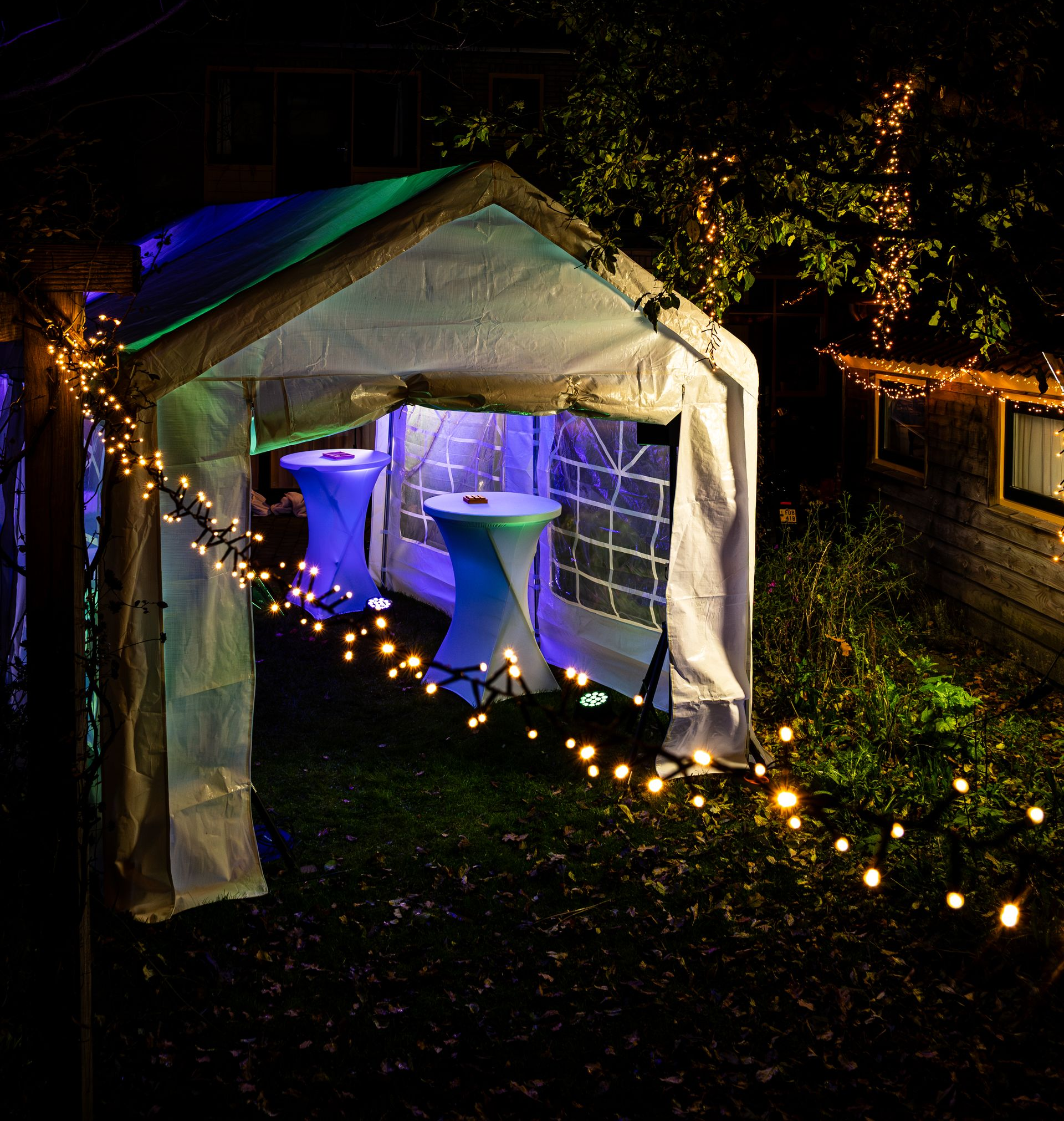 Een witte evenemententent voor buiten, 's nachts verlicht met een levendige blauwe gloed, staat op een grasveld te midden van donker gebladerte.