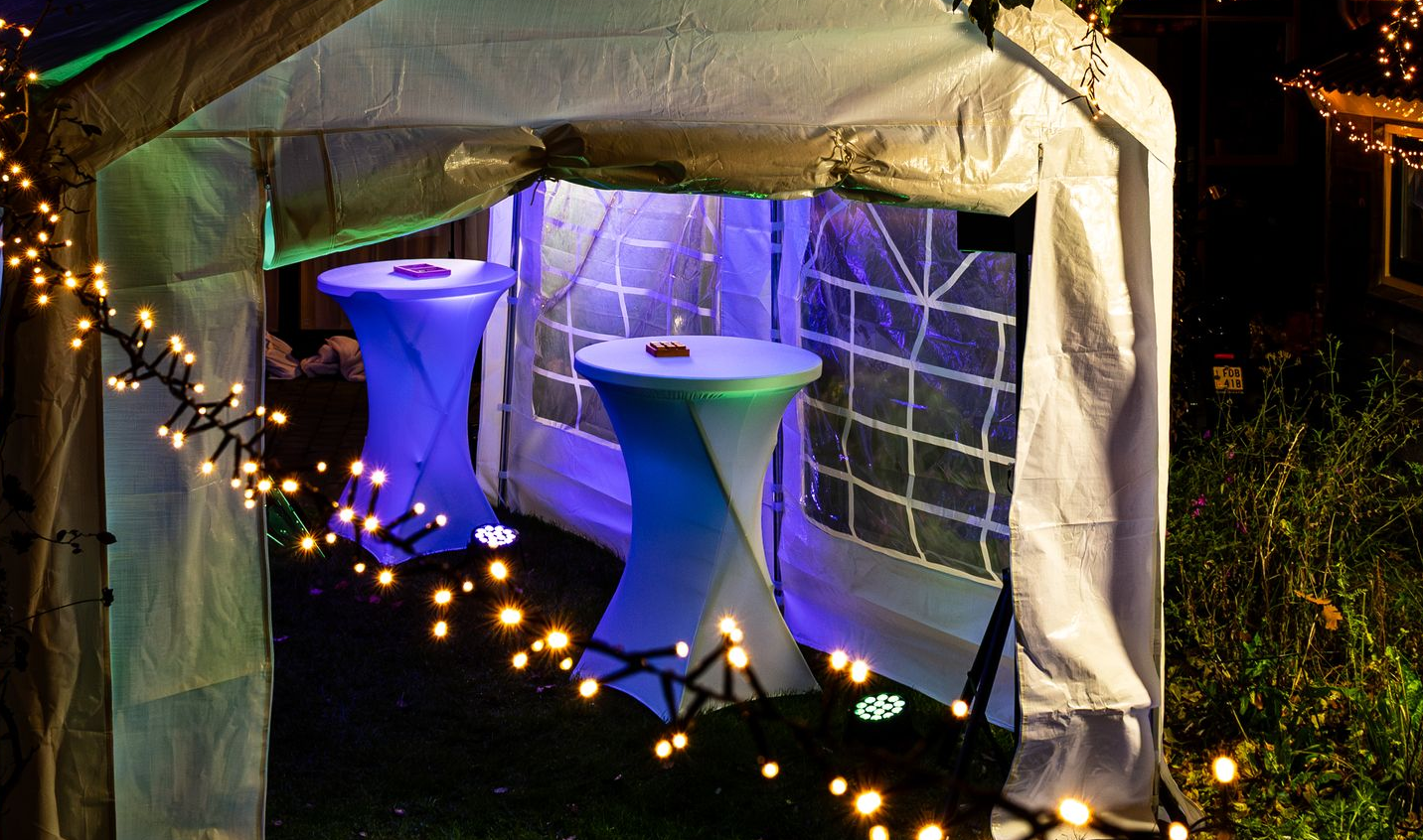 Twee lichtblauwe, glinsterende tafels op sokkels staan ​​in een witte feesttent die 's avonds verlicht wordt door warme lichtslingers.