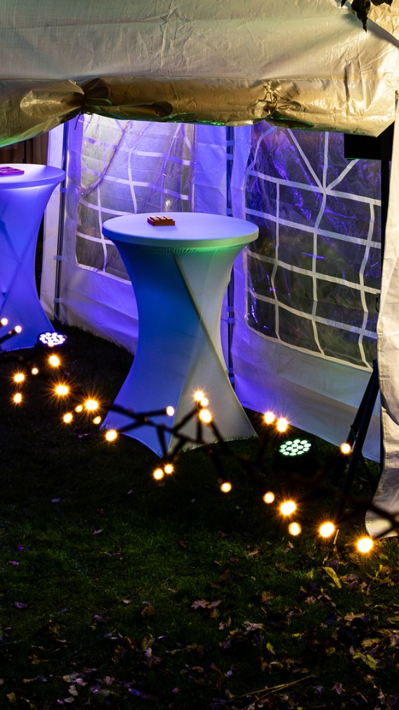 Twee lichtblauwe, glinsterende tafels op sokkels staan ​​in een witte feesttent die 's avonds verlicht wordt door warme lichtslingers.