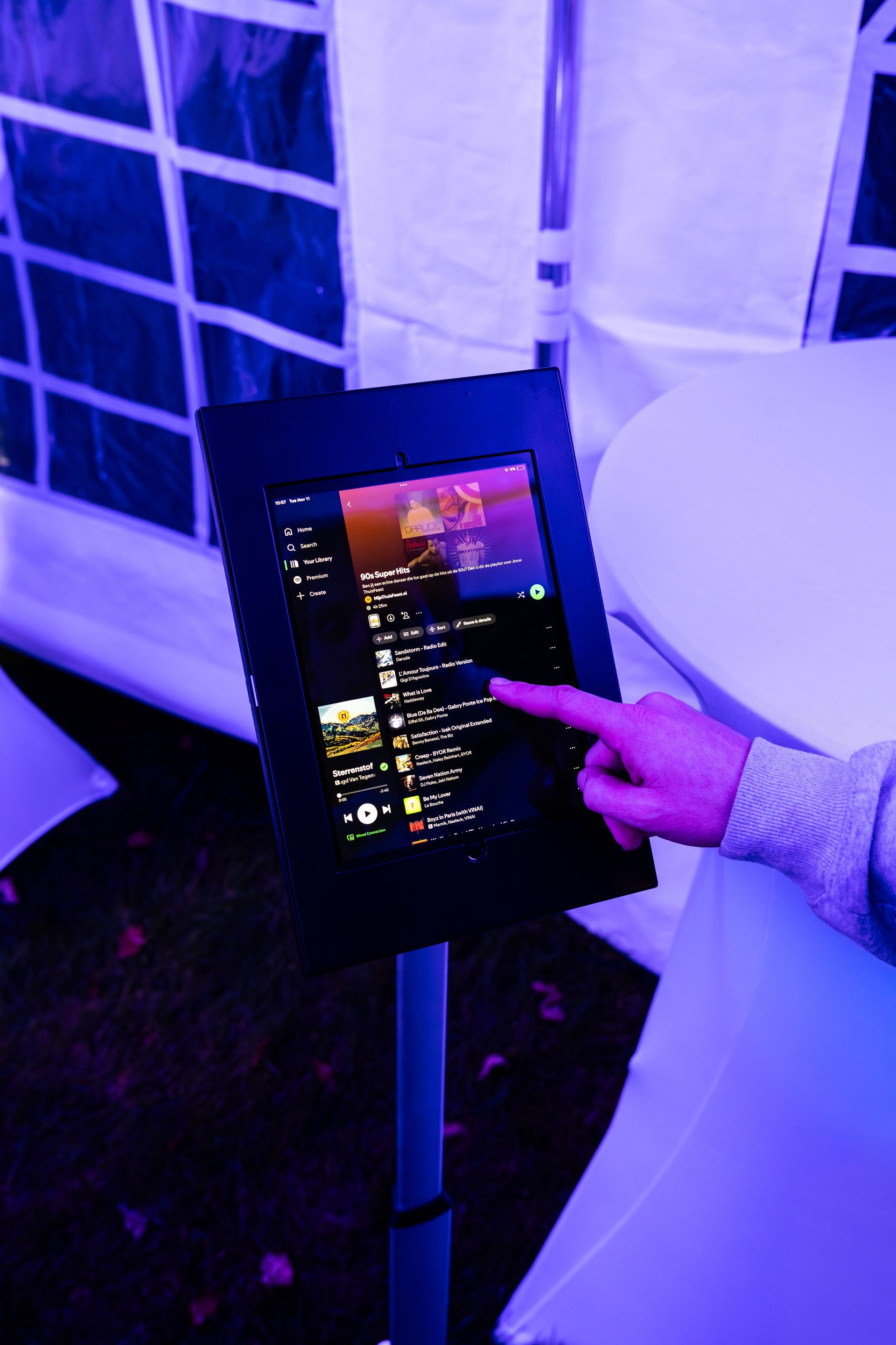 Een hand bedient een digitaal touchscreen dat op een standaard staat tijdens een buitenevenement dat in blauw licht is verlicht.