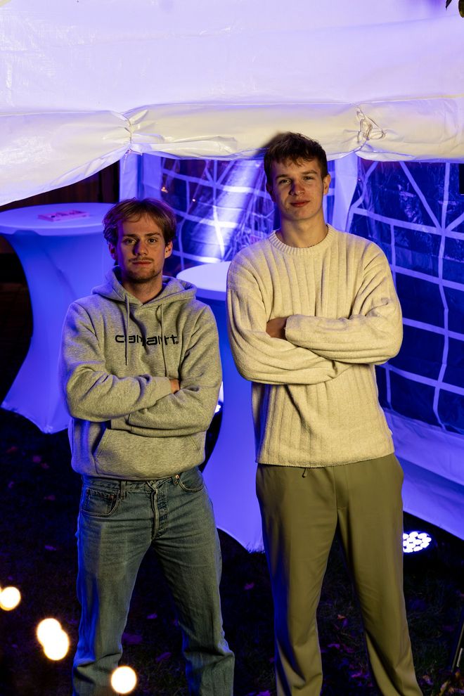 Twee jongemannen staan ​​voor een witte tent met blauwe verlichting. De een heeft zijn armen over elkaar, de ander zijn handen in zijn zakken.