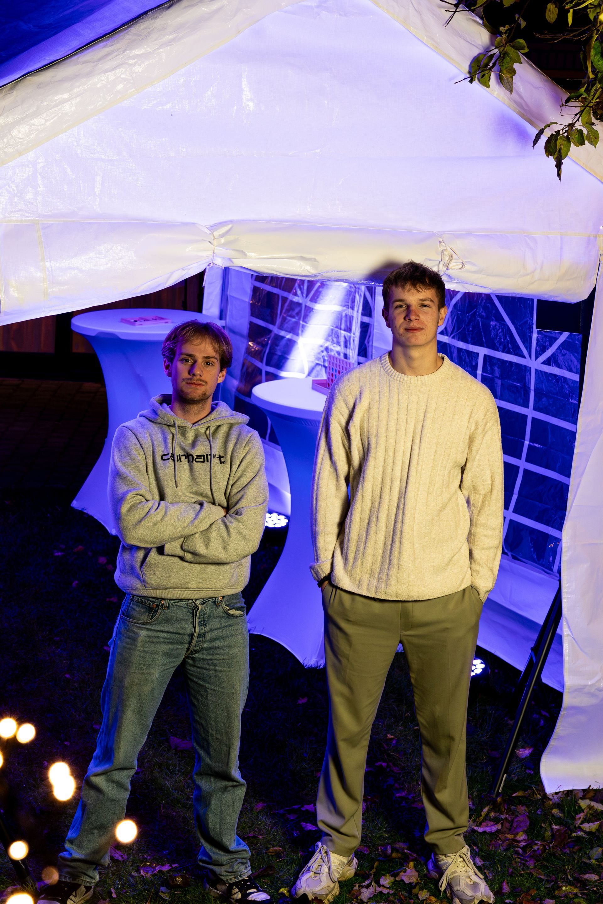 Twee mensen staan ​​onder een witte tent die verlicht wordt door blauw licht; de een draagt ​​een grijze hoodie, de ander een crèmekleurige trui.