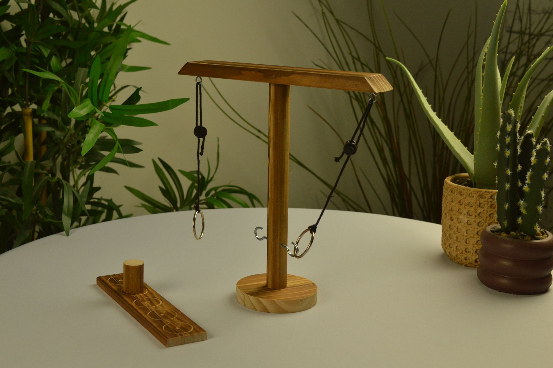 Een houten ringwerpspel met twee hangende ringen op een standaard staat op een witte tafel naast kamerplanten.