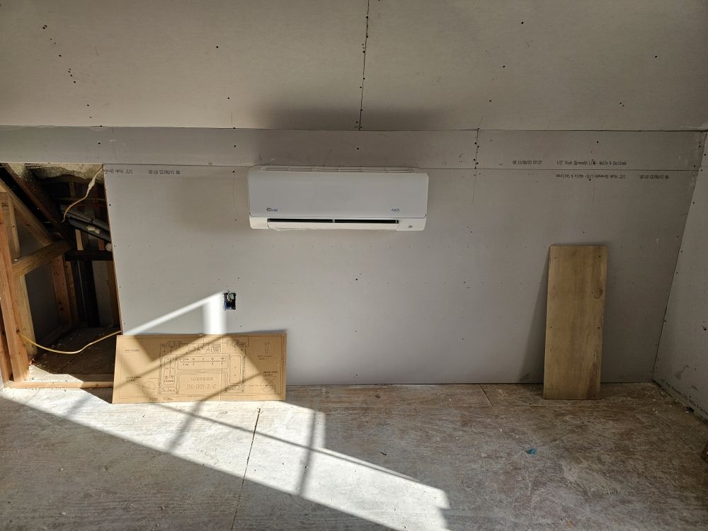 A room under construction with an air conditioning unit mounted on a gray wall. Sunlight streams across the floor.