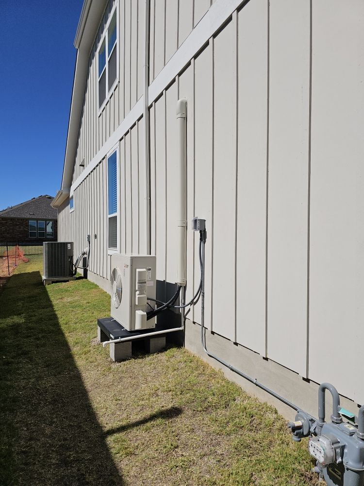 An outdoor air conditioning unit is mounted on a concrete block next to a building with vertical siding. The sky is blue.