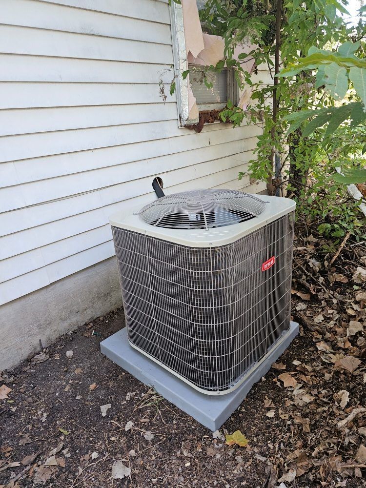 Air conditioning unit on a concrete pad next to a white house. Green foliage surrounds it.