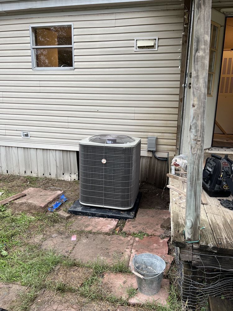 An air conditioning unit sits on a black base next to a building with beige siding. A window and electrical box are visible.