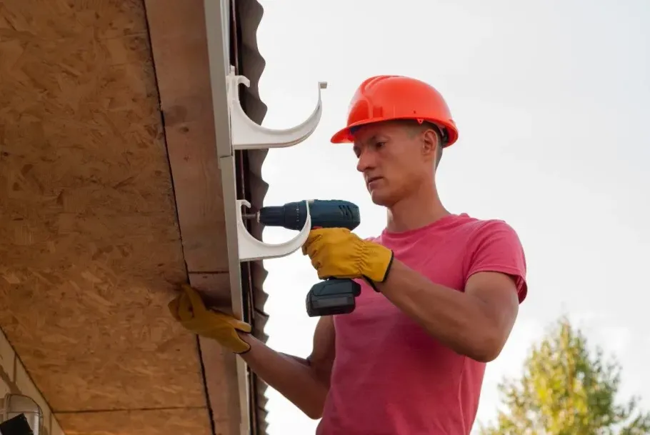 Worker in orange hard hat using a drill to install gutters on a house roofline
