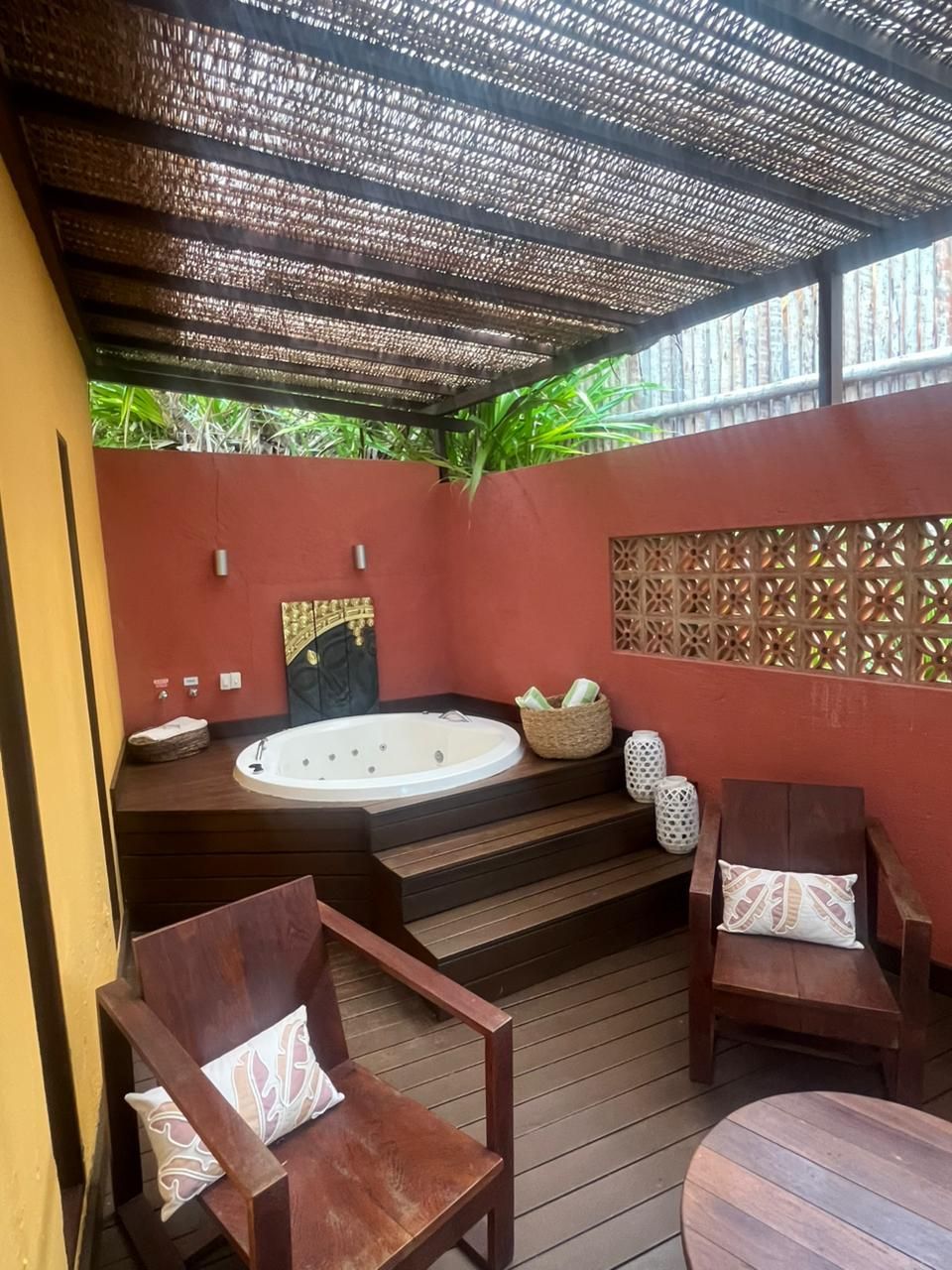 Área de spa ao ar livre com banheira de hidromassagem, cadeiras e teto texturizado. Paredes em tons de terra e deck de madeira.