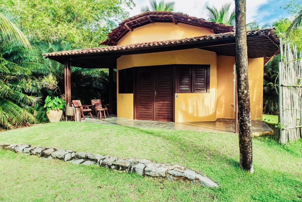 Bangalô amarelo com telhado marrom e móveis na varanda, situado em um quintal gramado com árvores.