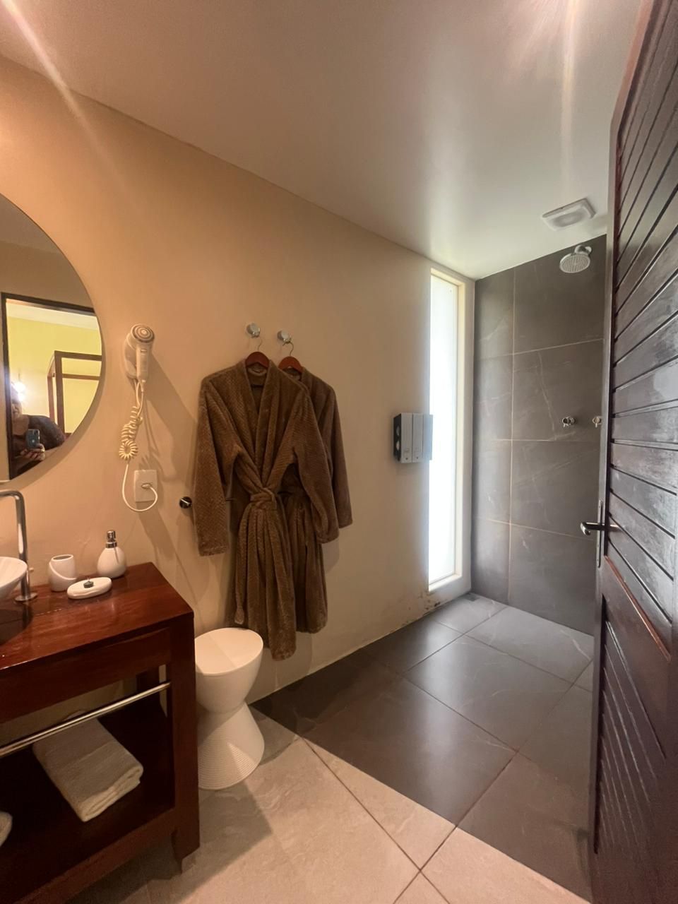 Banheiro com chuveiro, roupões e penteadeira. Paredes bege, azulejos cinza, madeira marrom.