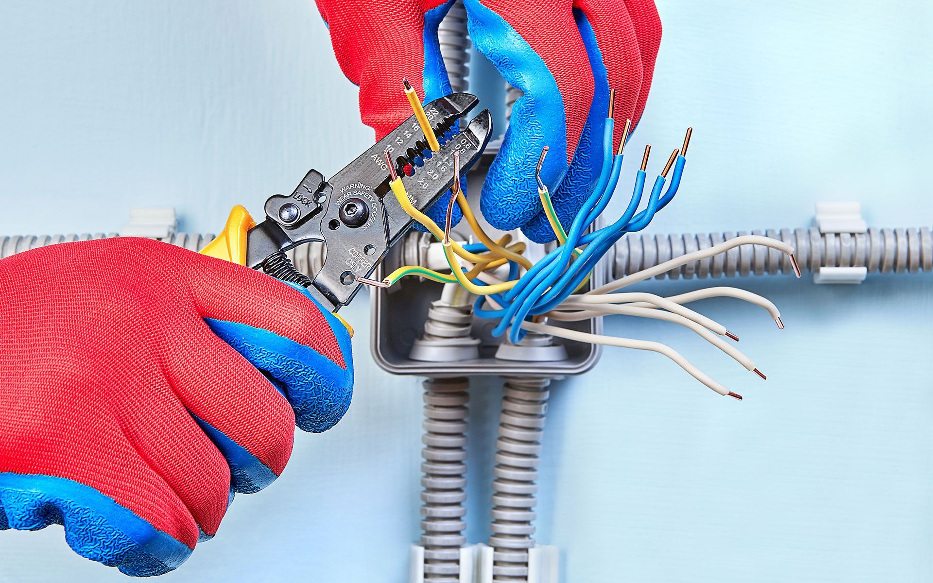 Electrician using wire strippers on electrical wires in a wall-mounted junction box.